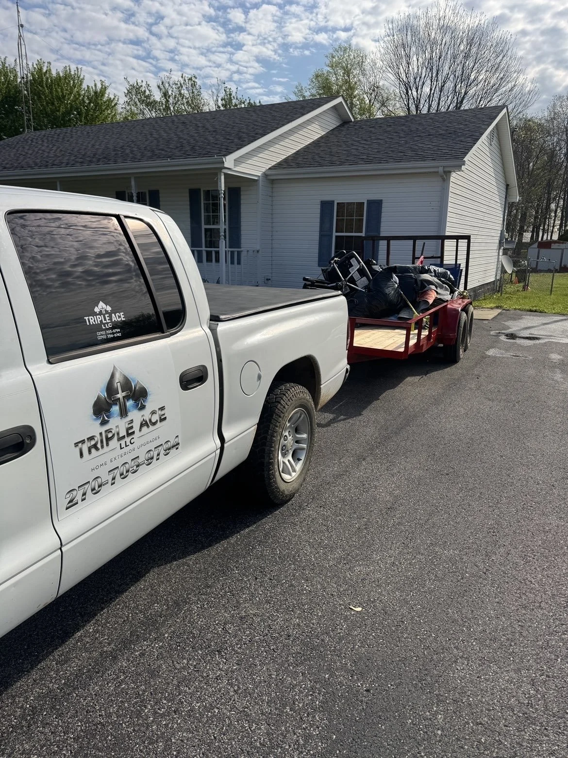 A pickup truck with a logo and contact number for Triple Ace LLC parked in a driveway, attached to a red enclosed trailer loaded with black garbage bags and miscellaneous items, in front of a white house with blue shutters.