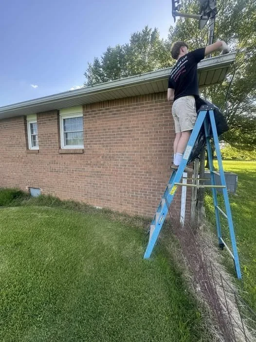 Young man on a blue ladder fixing or installing a satellite dish on the side of a brick house in a grassy yard.