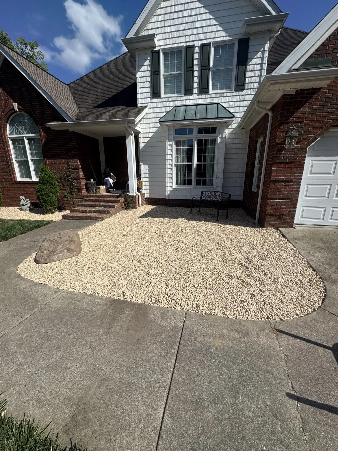 Front yard of a house featuring a gravel landscaping area with a large rock, concrete sidewalk, house with a brick and white siding exterior, multiple windows, a small porch with stairs, a bench, and a person sitting on the porch.