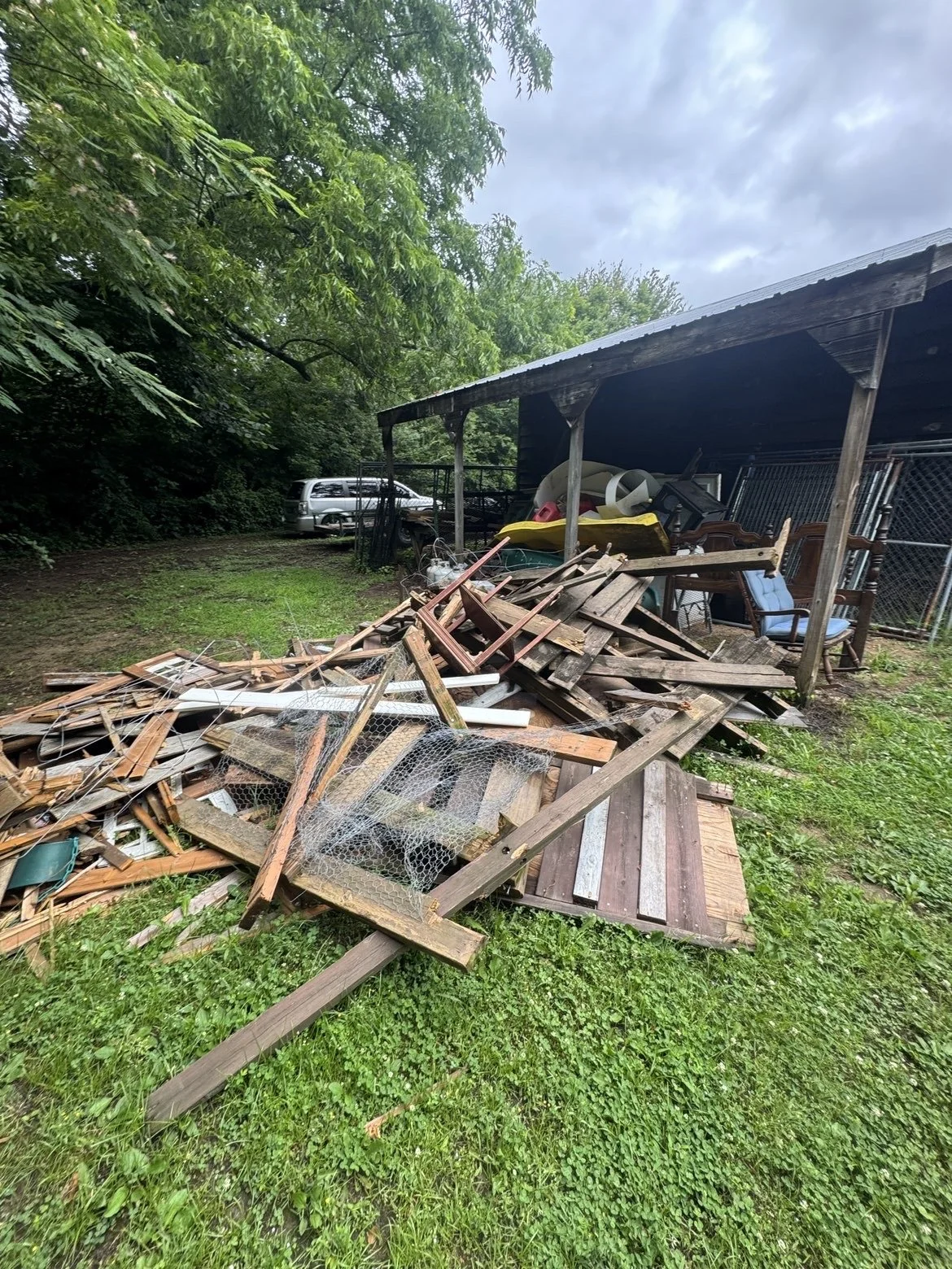 A pile of discarded wooden debris and various household items on grass in front of a wooden shed with chairs and outdoor equipment, with trees and a cloudy sky in the background.