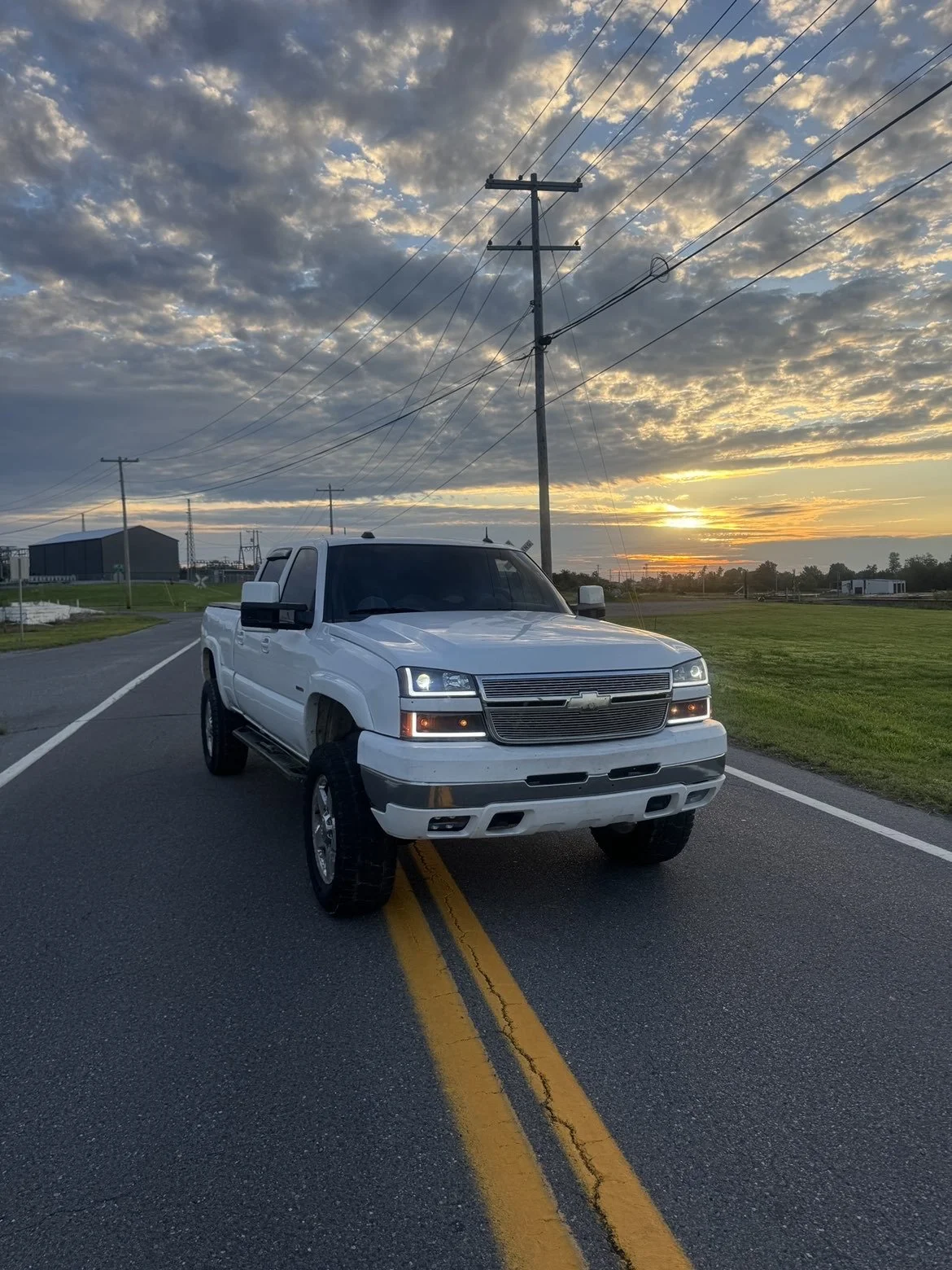 A white Chevrolet pickup truck parked on a two-lane road at sunset, with power lines overhead and a grassy field on the side.