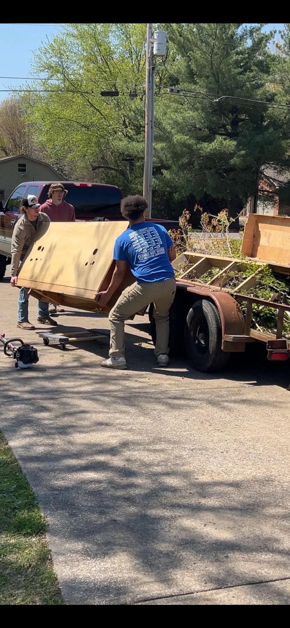 Three young men loading a large wooden object onto a flatbed trailer parked on a residential street, with trees and a power pole in the background.