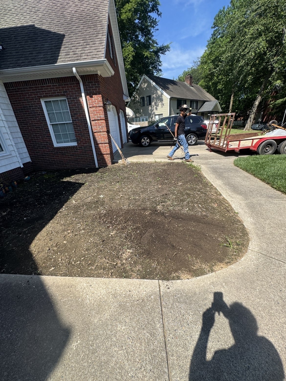 A man working on a garden bed in a front yard, with a house, driveway, and parked cars in the background, on a sunny day.
