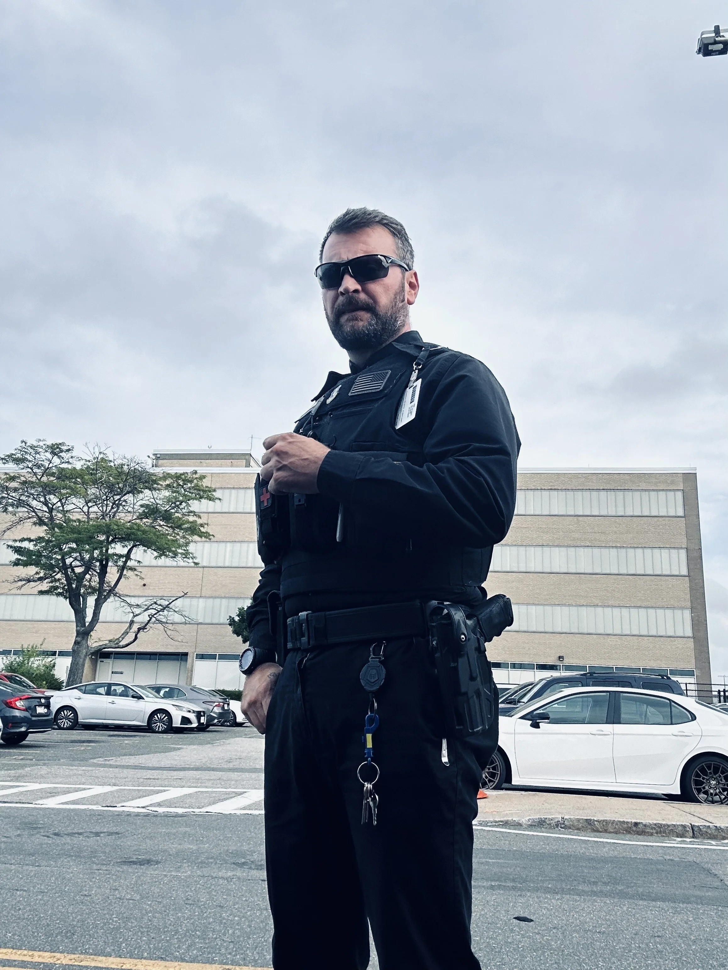 A police officer standing in a parking lot, wearing black uniform and sunglasses, with a multi-story building and parked cars in the background.