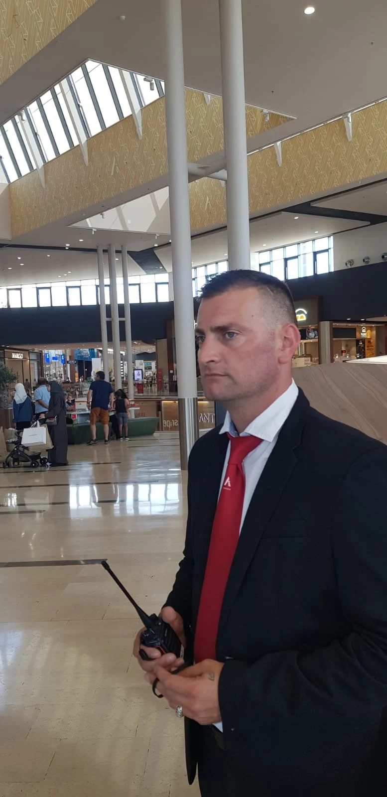 A man in a business suit with a red tie holding a walkie-talkie at an airport terminal.