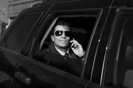 A man in a suit and sunglasses talking on a mobile phone inside a black car.