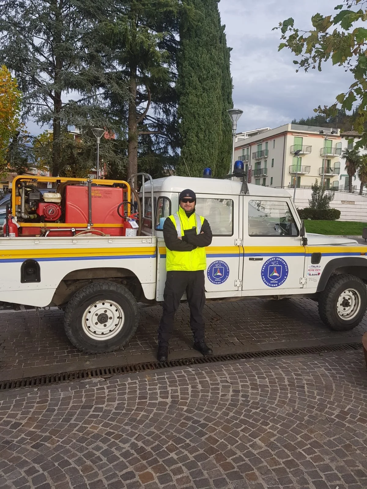 A man in a bright yellow safety vest and black clothing, wearing sunglasses and a beanie, standing with arms crossed next to a white emergency vehicle with blue markings and emergency lights on top, parked on a cobblestone street with tall trees and apartment buildings in the background.