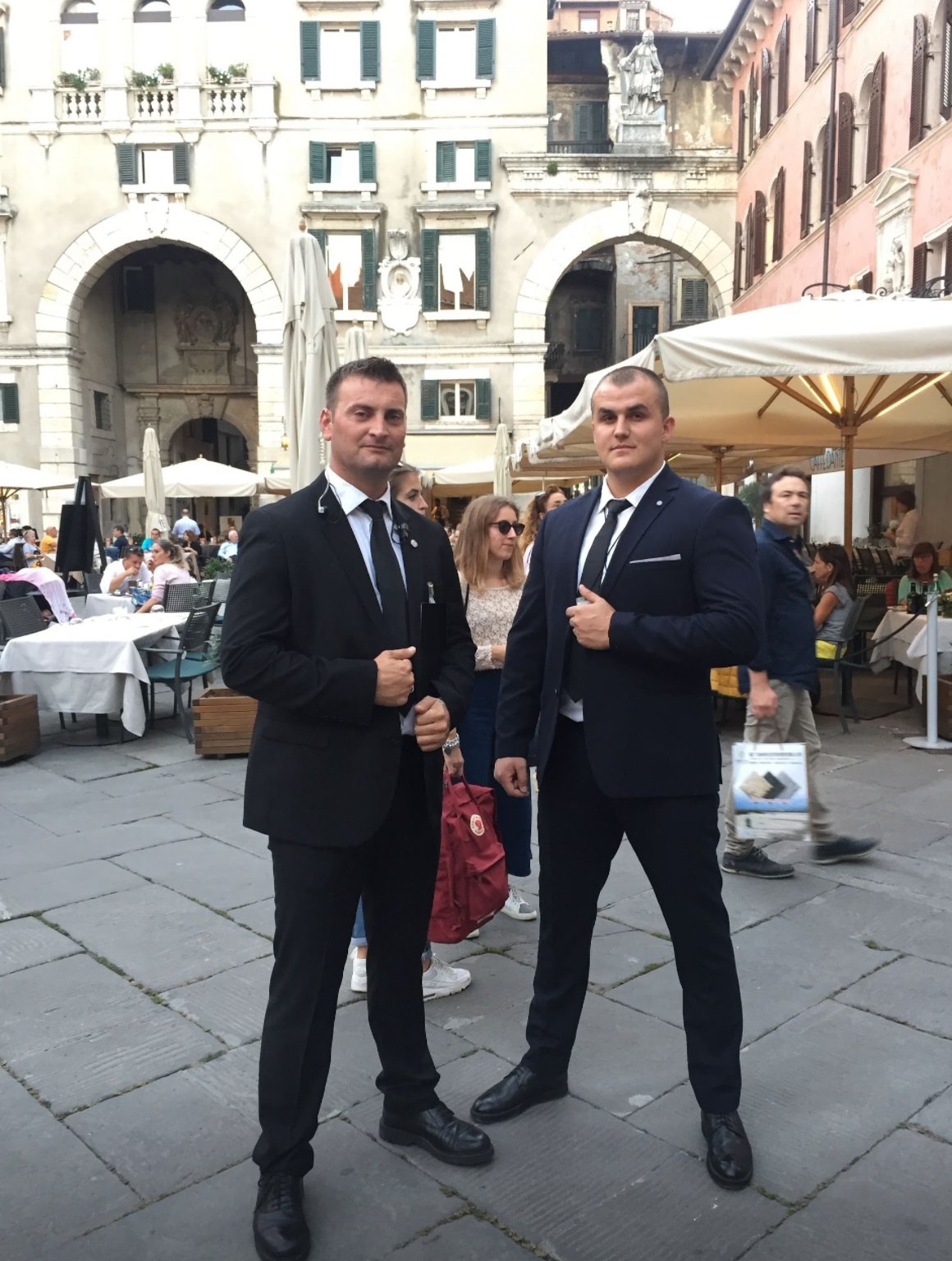 Two men in dark suits and ties standing on a cobblestone street in a European city, with outdoor restaurant seating and historic buildings in the background.