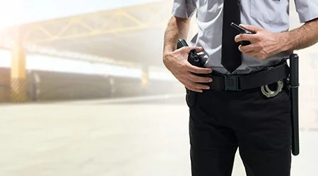 Close-up of a police officer's waist holding a walkie-talkie and a phone, with an industrial or construction site background.