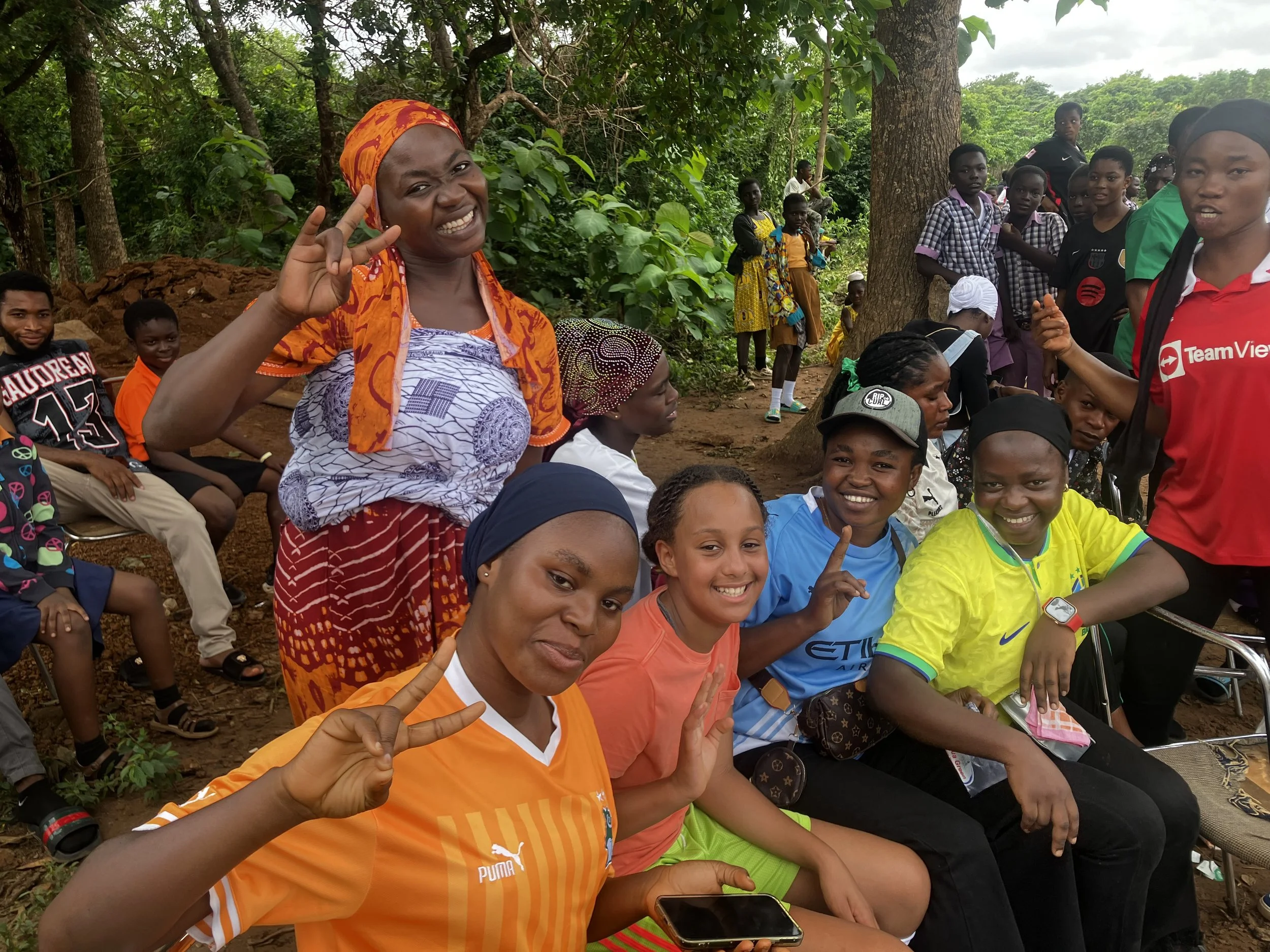 Group of smiling women and children outdoors, some making peace signs and gestures, in a forested area.