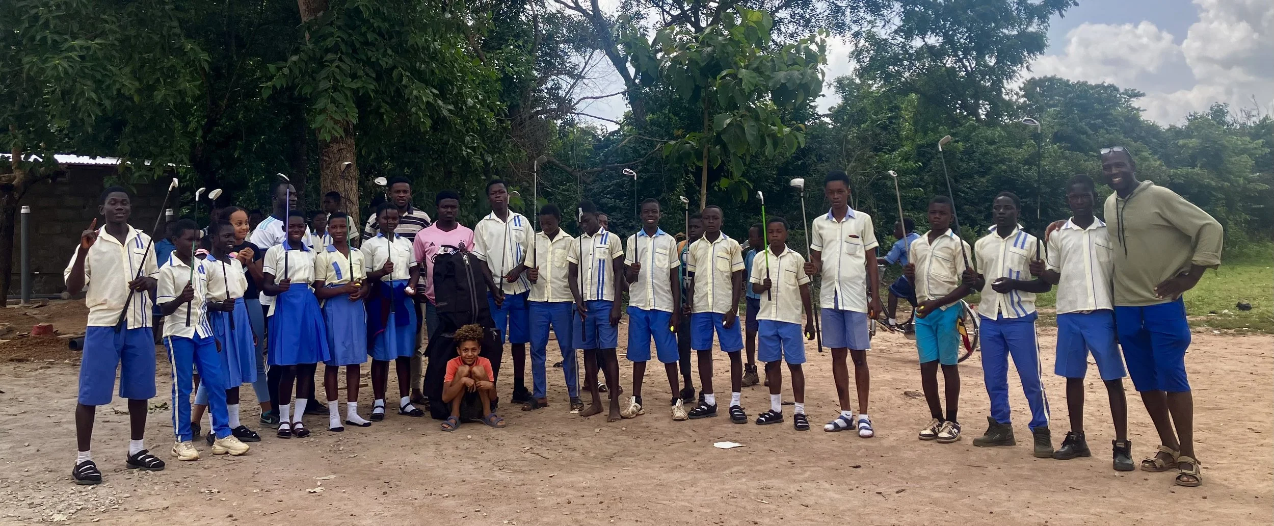 Group of students in school uniforms standing outdoors, holding golf clubs, with a teacher or coach on the right, under a partly cloudy sky.