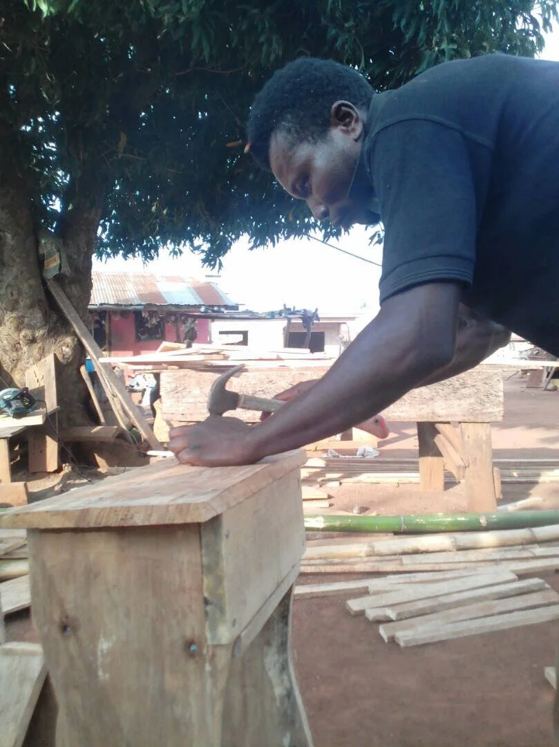 Man working on a piece of wood with a hammer in an outdoor woodworking shop, under a large tree, with various wooden materials and tools around.