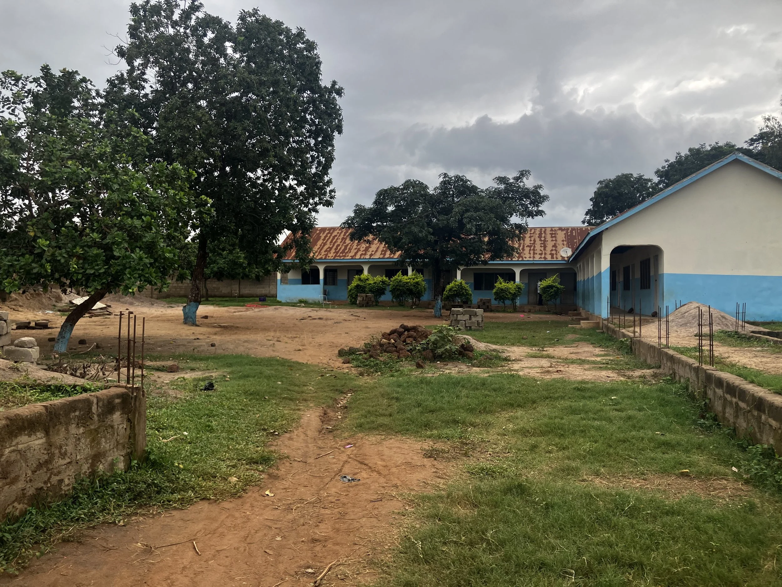 A school building with a rusty roofing, surrounded by trees and a partially grassy yard under cloudy skies.