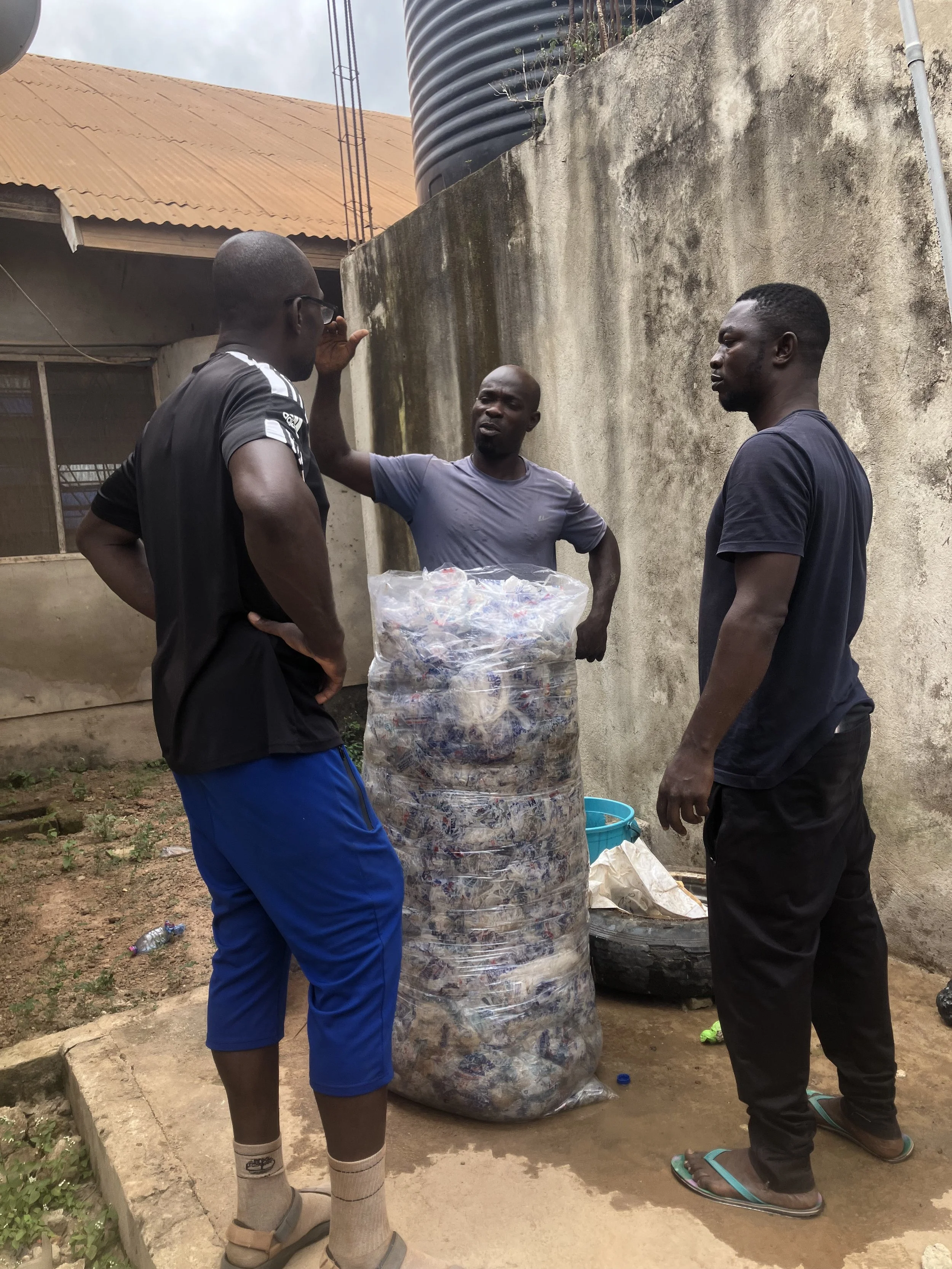 Three men standing outdoors around a large bag of crushed plastic bottles, engaging in conversation. One man is speaking and gesturing with his hand. The setting appears to be a backyard or alley with an old wall and water tank in the background.