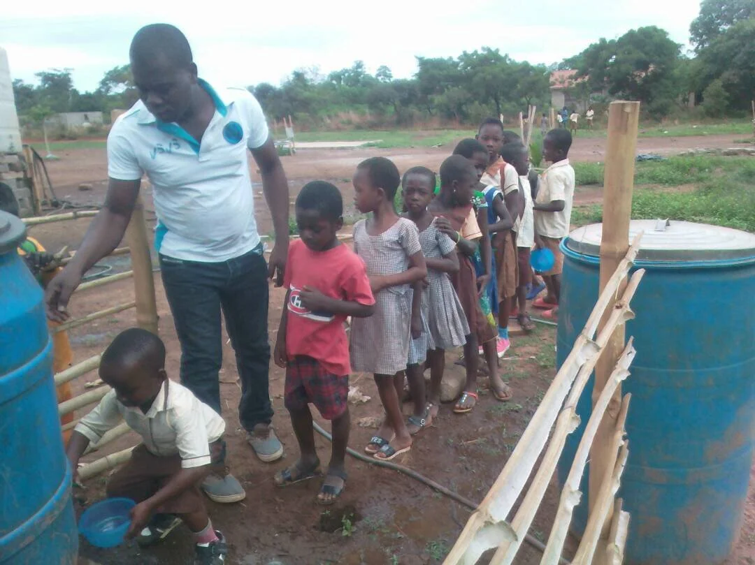 A group of children standing in line outdoors, with an adult supervising near blue water containers and bamboo structures.