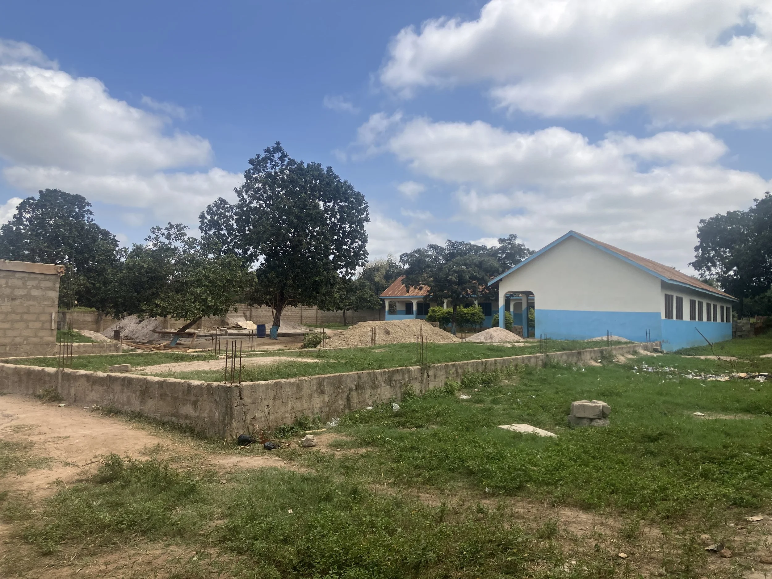 A rural scene with a single-story white and blue building, trees, patches of grass, and a dirt pathway, under a partly cloudy sky.