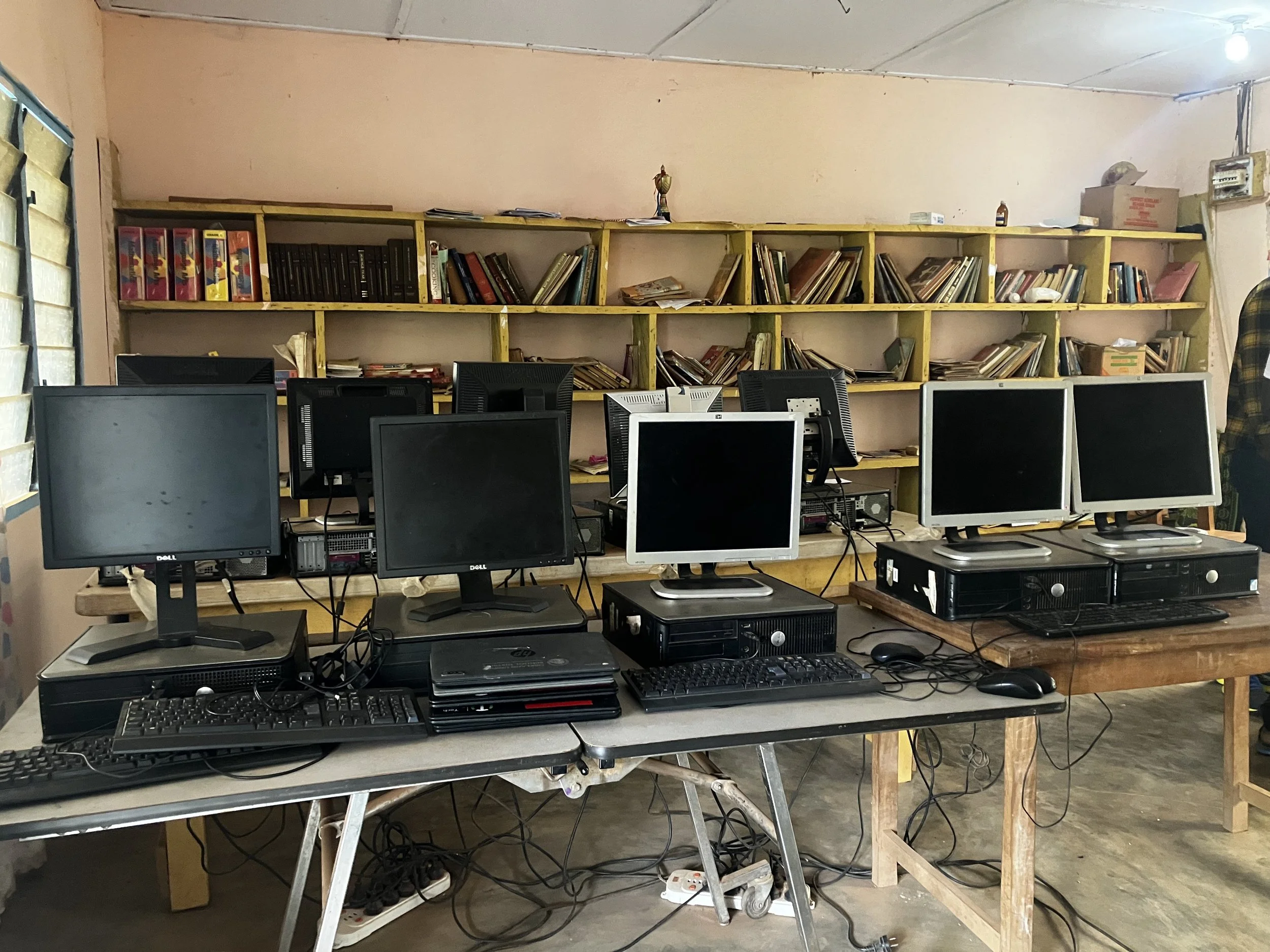 A room with a wooden table holding multiple desktop computers, monitors, keyboards, and mice, with a yellow shelf on the wall in the background filled with books and files.