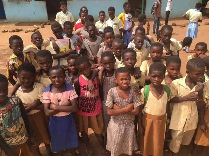Group of school children standing outdoors, some with backpacks, posing for a photo with buildings in the background.