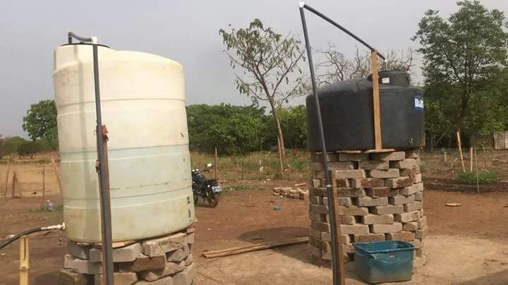 Two water containers, one white and one black, elevated on stacks of bricks outdoors with trees and open land in the background.