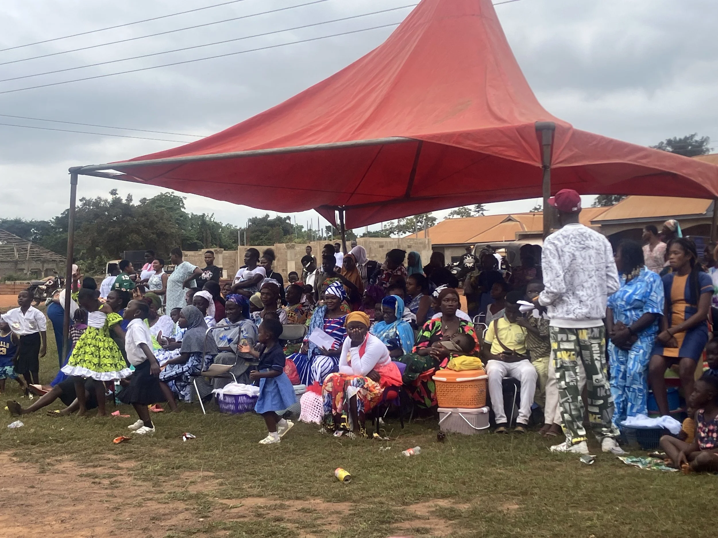 A large gathering of people, including children and adults, sitting and standing under a red canopy outdoors on a grassy field, with some buildings and trees in the background, during a daytime event.