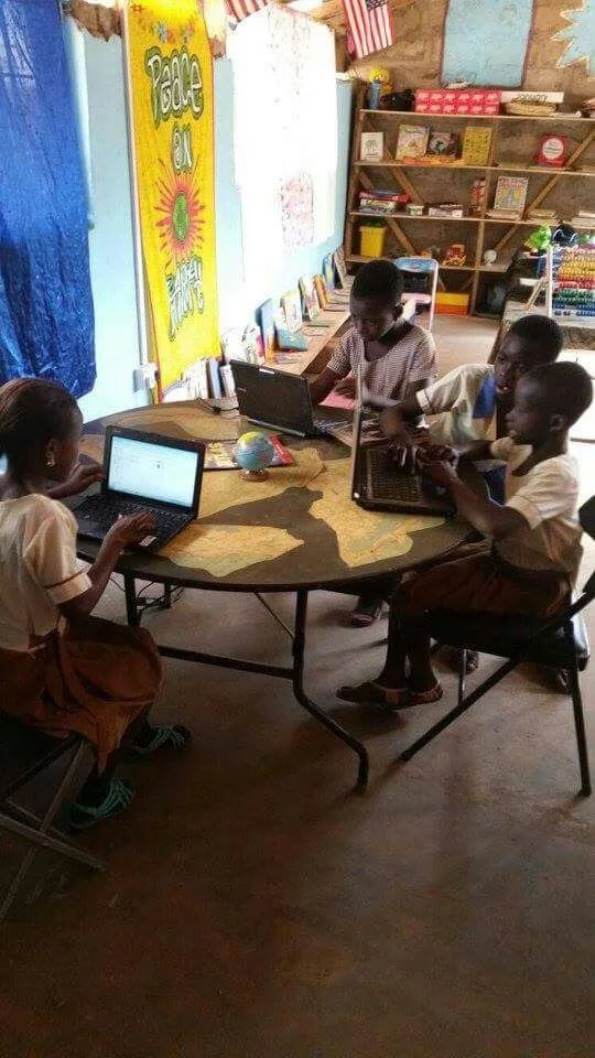 Four children sitting around a table using laptops in a classroom with bookshelves and educational posters.