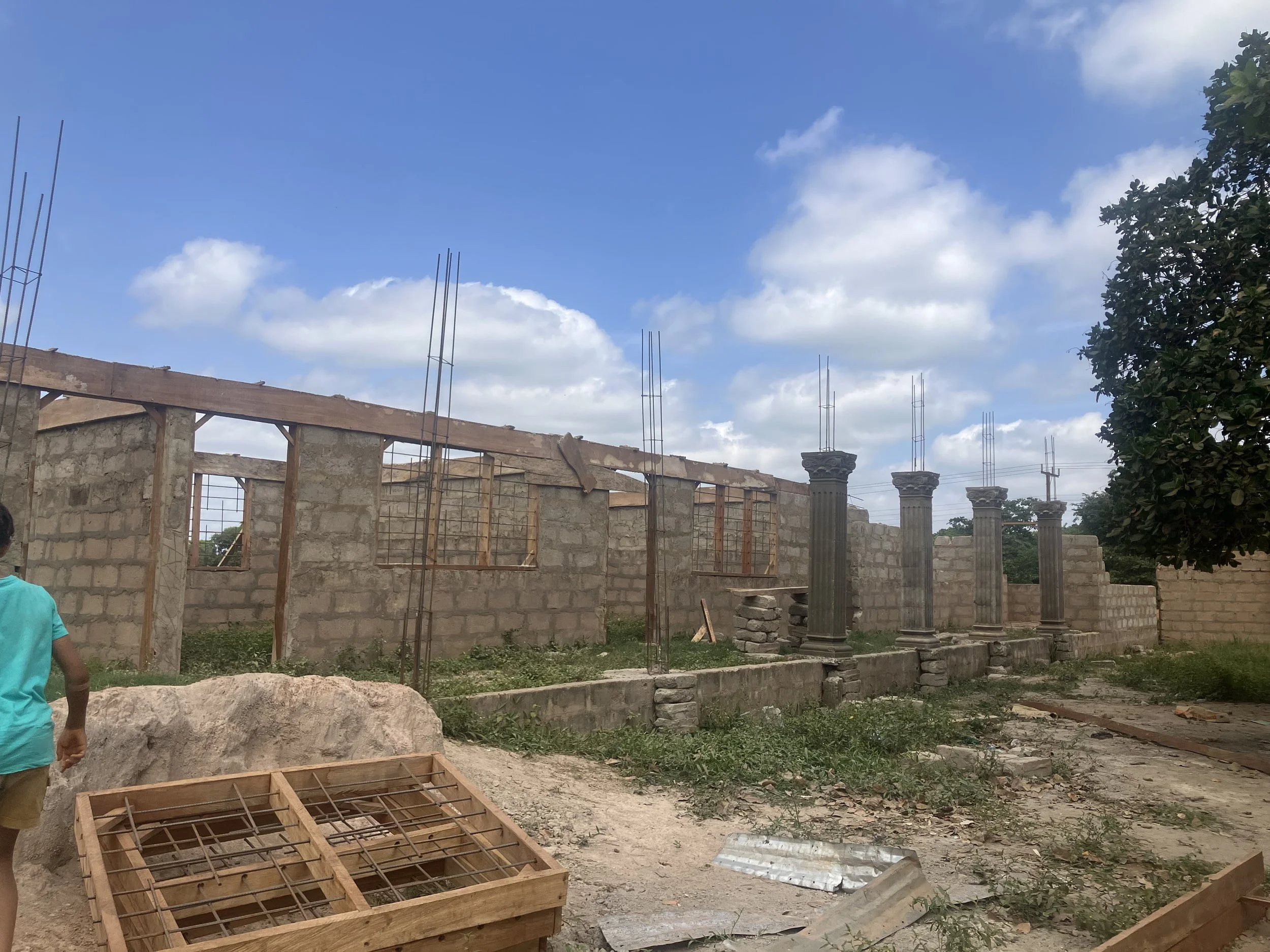 Construction site with partially built stone and brick walls, four columns with decorative capitals, and construction materials on ground about a sunny day with blue sky and scattered clouds.