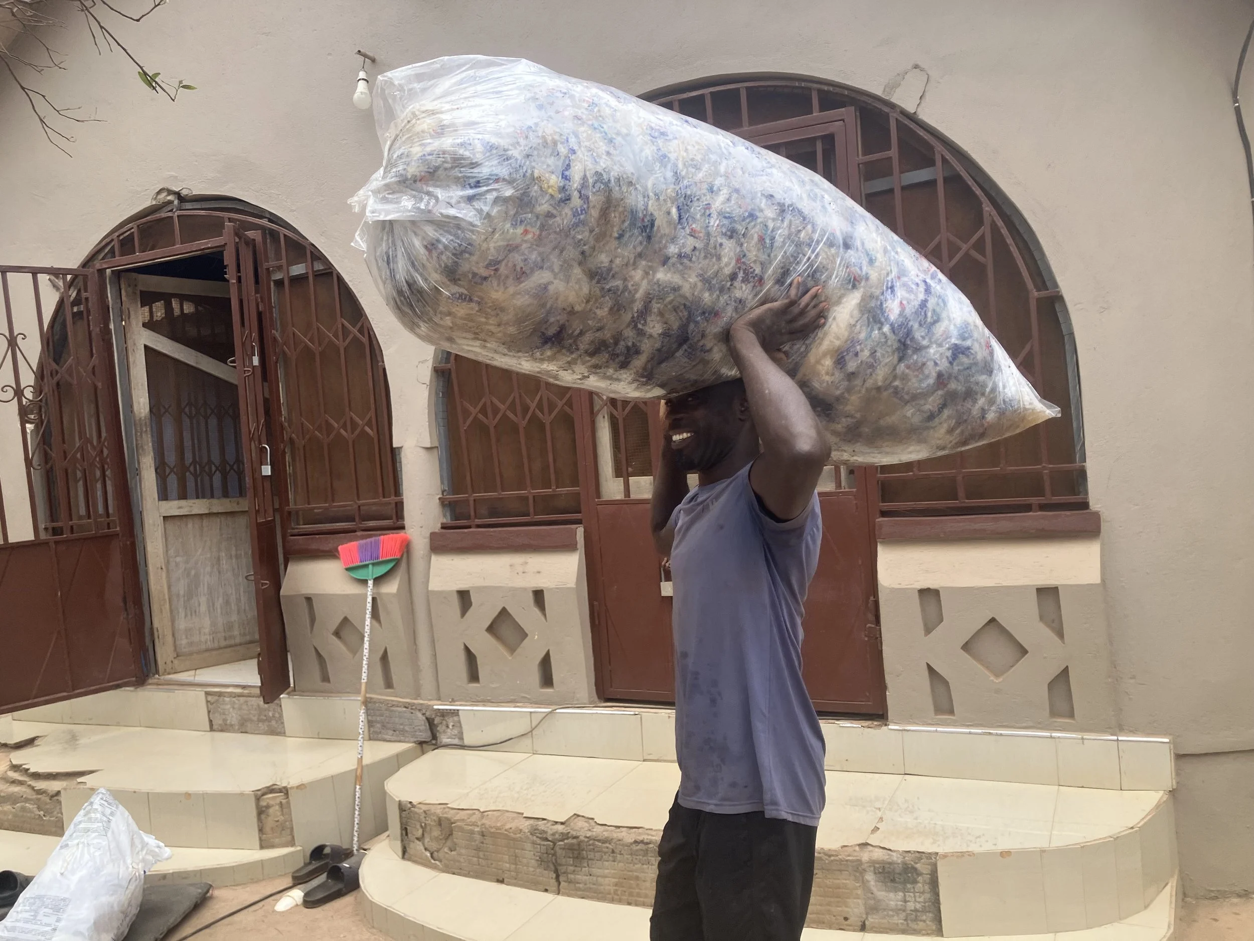 A man smiling, balancing a large plastic bag filled with colorful items on his shoulder, standing outside a building with arched windows and a tiled staircase.