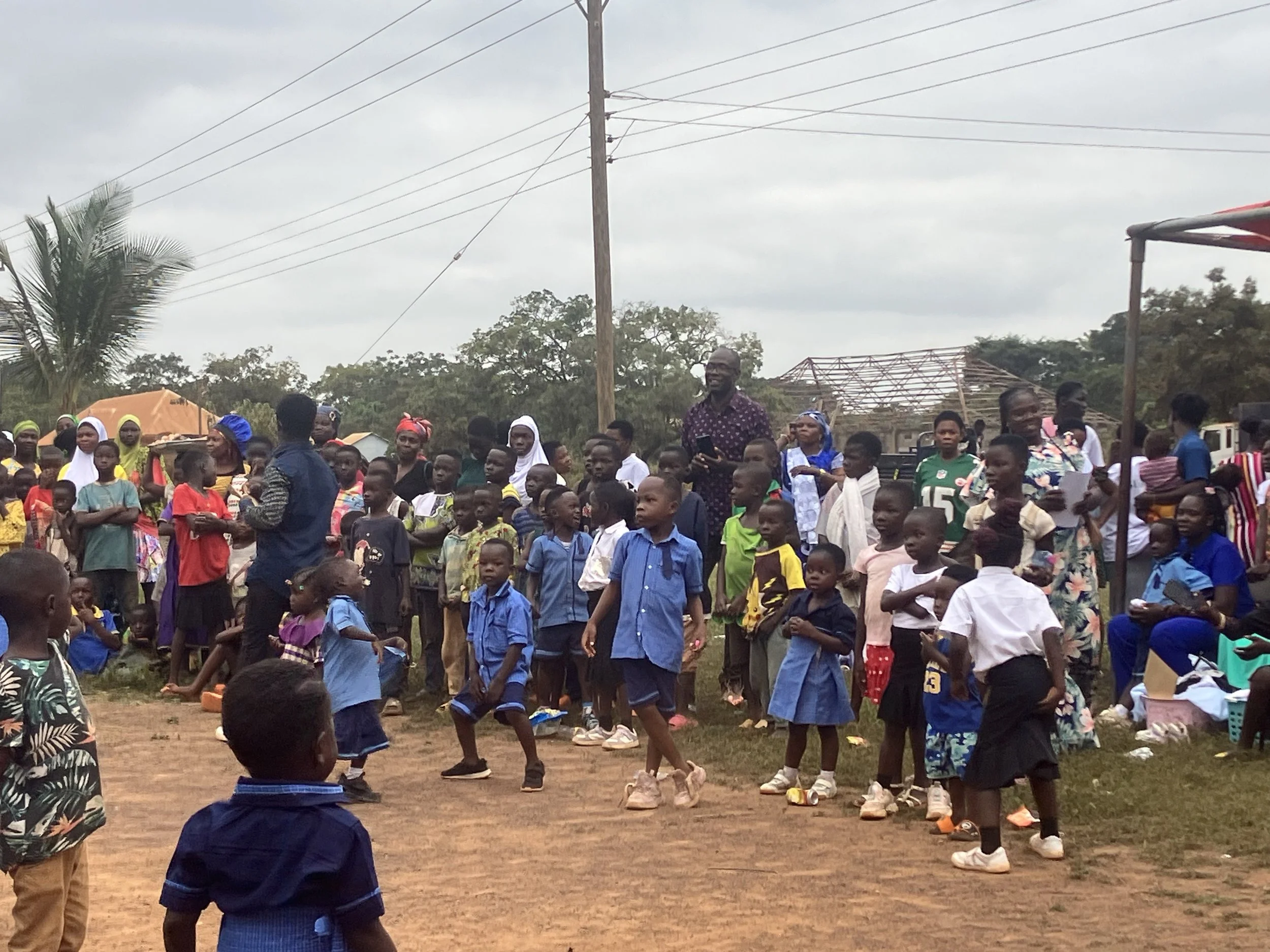 A large group of children and adults gathered outdoors on a cloudy day, some standing and some sitting, with a few young children dancing on a dirt ground. There are houses and trees in the background.