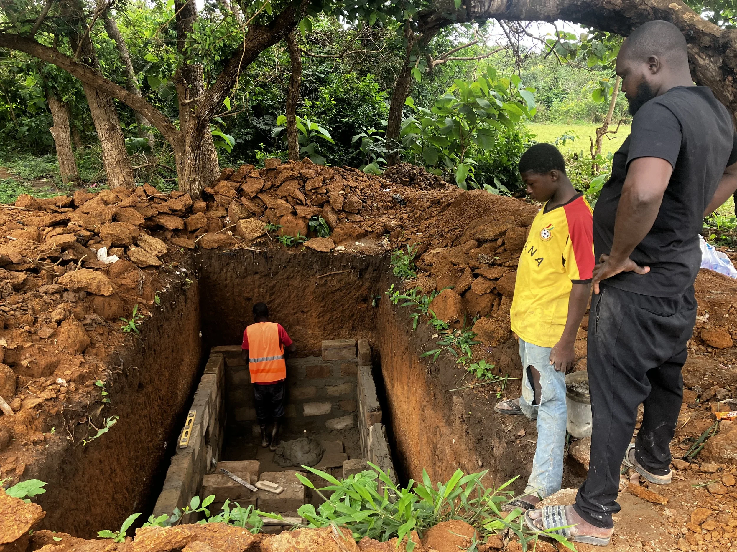 Two men and a boy stand at the edge of a construction site in a wooded area, digging and building a rectangular structure with bricks and concrete.