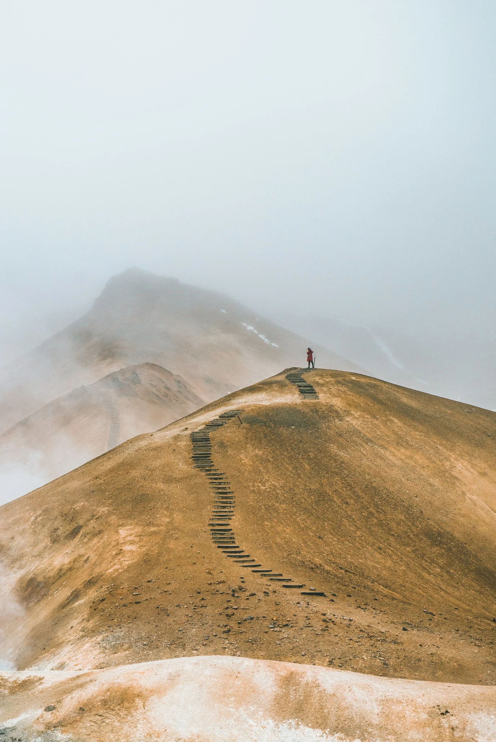A person standing on a mountain ridge with a winding staircase leading to the top, surrounded by mist and barren landscape.