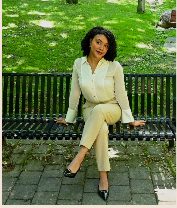 A woman in a cream-colored pantsuit sitting on a black bench outdoors in a park with green grass and trees in the background.