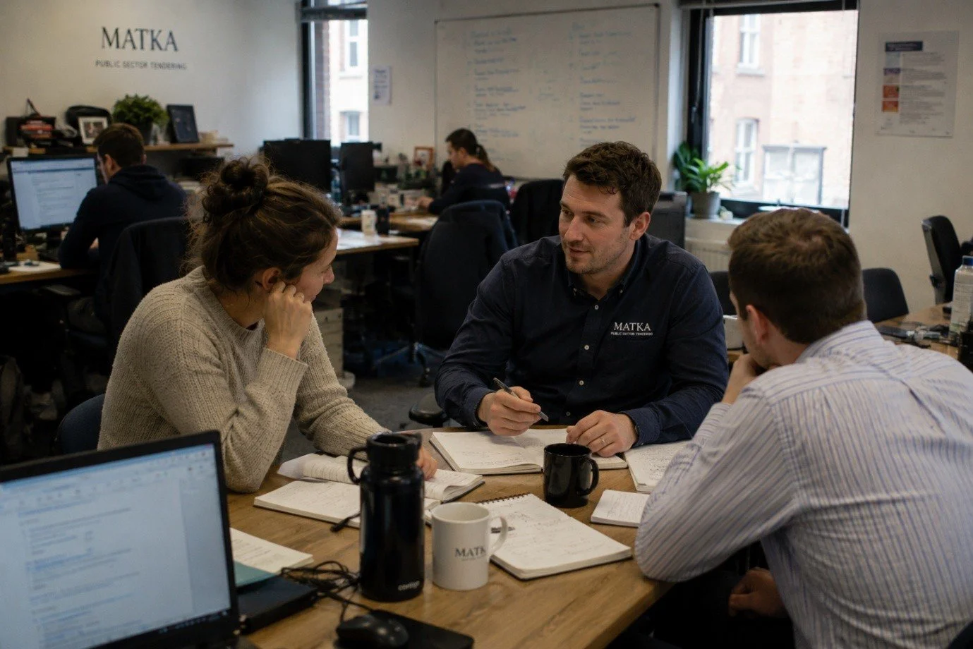 People in a meeting or discussion at an office, with laptops and notebooks on the table, in a workspace with windows and desks.