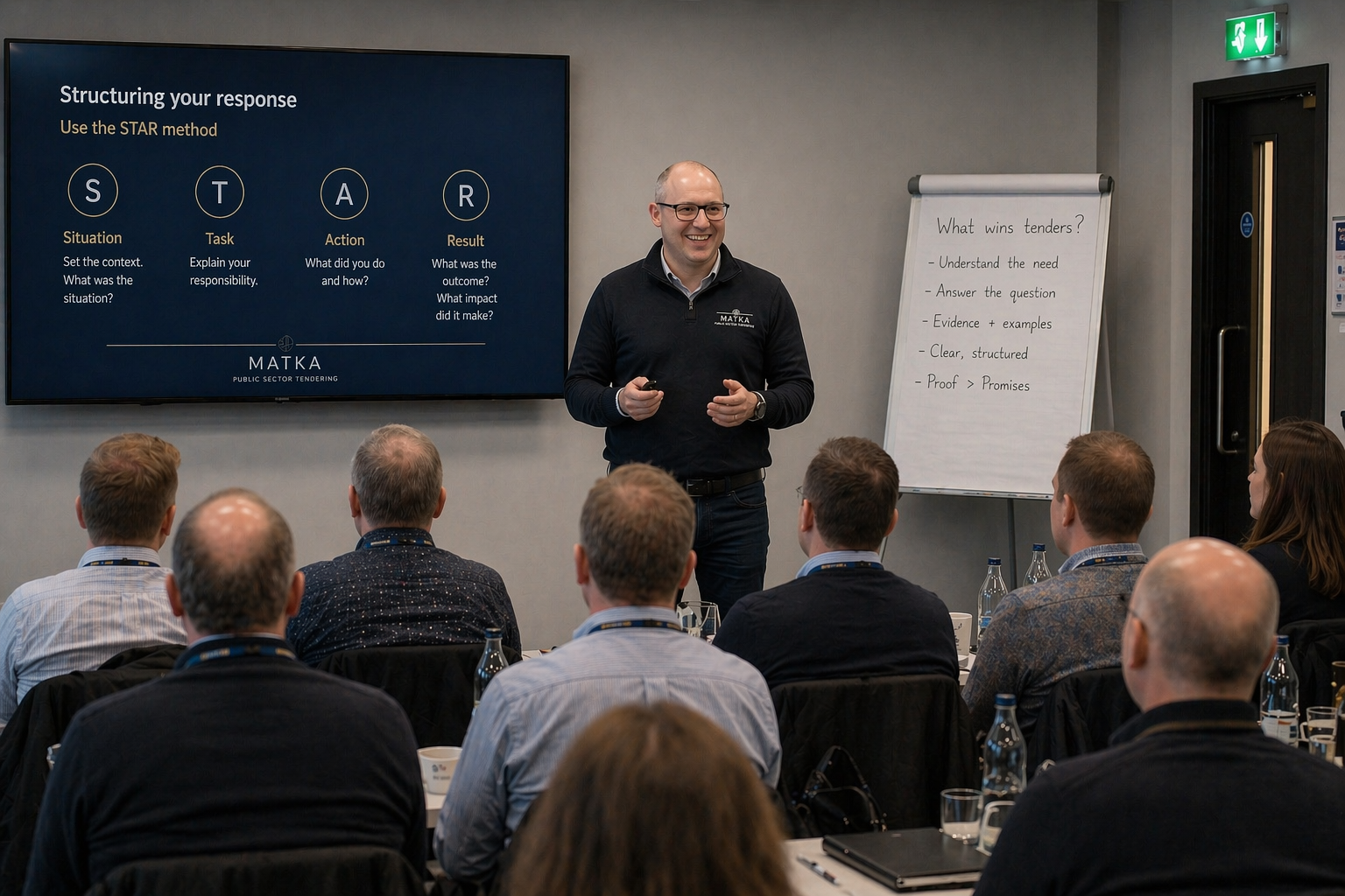 A man giving a presentation in a conference room with a large screen displaying the 'STAR' method for structuring responses. Several attendees are seated, listening attentively. There is also a whiteboard with notes on tenders and a green emergency exit sign above a door.