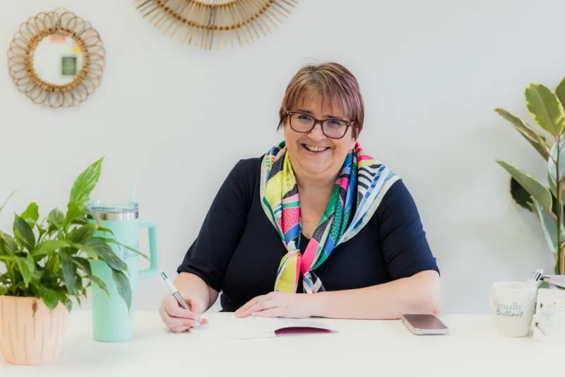 Smiling woman with short hair and glasses sitting at a desk with plants, a smartphone, and a notebook.