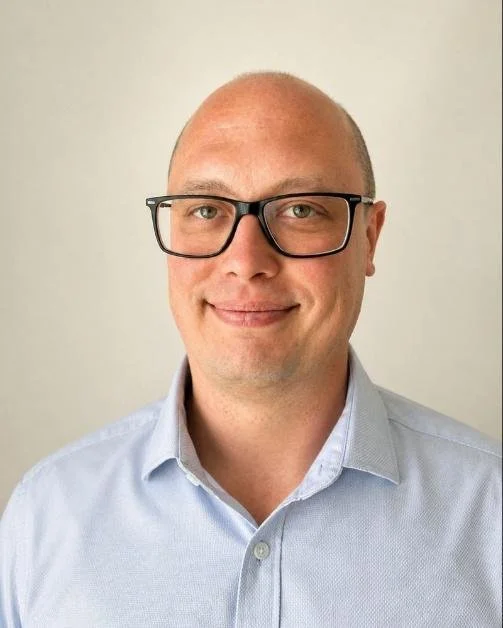 A bald man wearing black-rimmed glasses and a light blue button-up shirt, smiling at the camera against a plain light background.