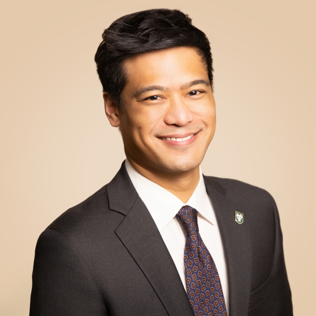 A young man with dark hair, wearing a dark suit, white shirt, and patterned tie, smiling against a plain beige background.