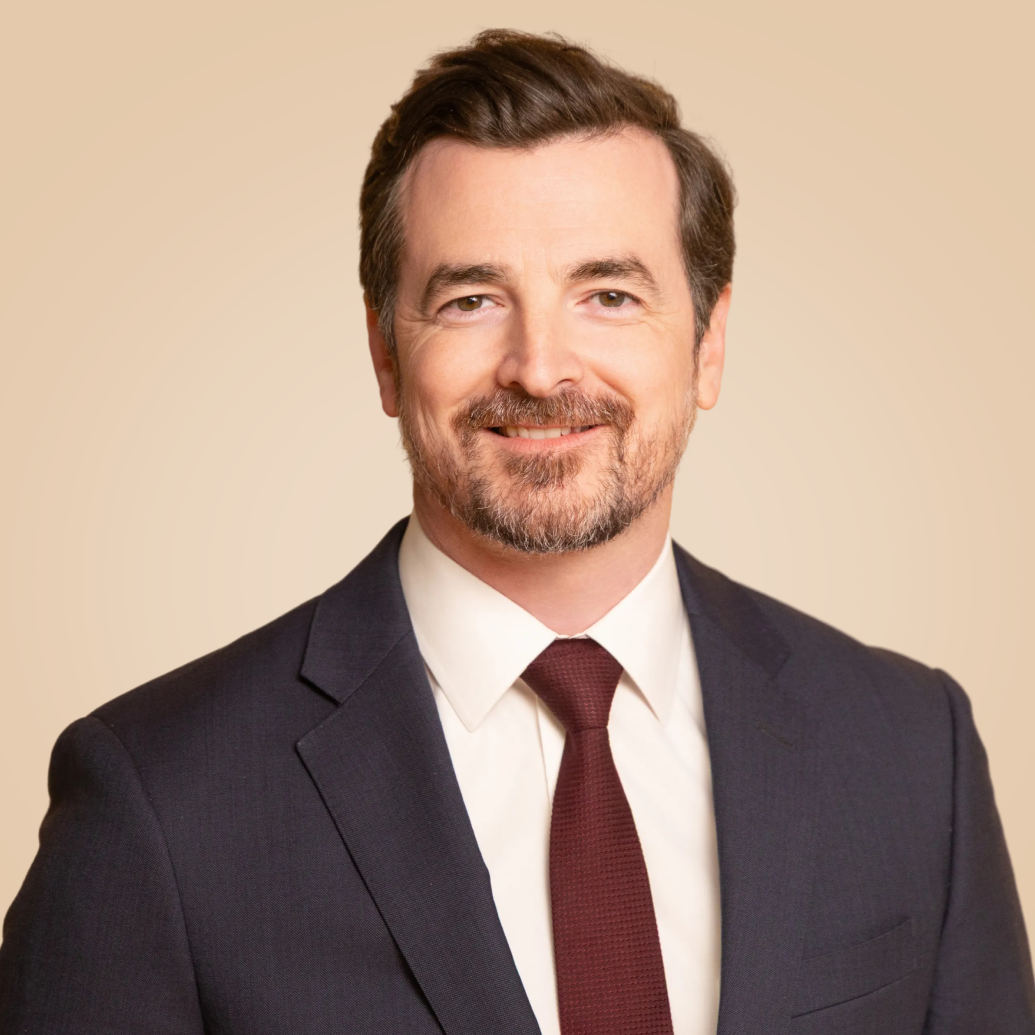 A professional man with brown hair, beard, and mustache, wearing a dark suit, white dress shirt, and maroon tie, smiling against a beige background.