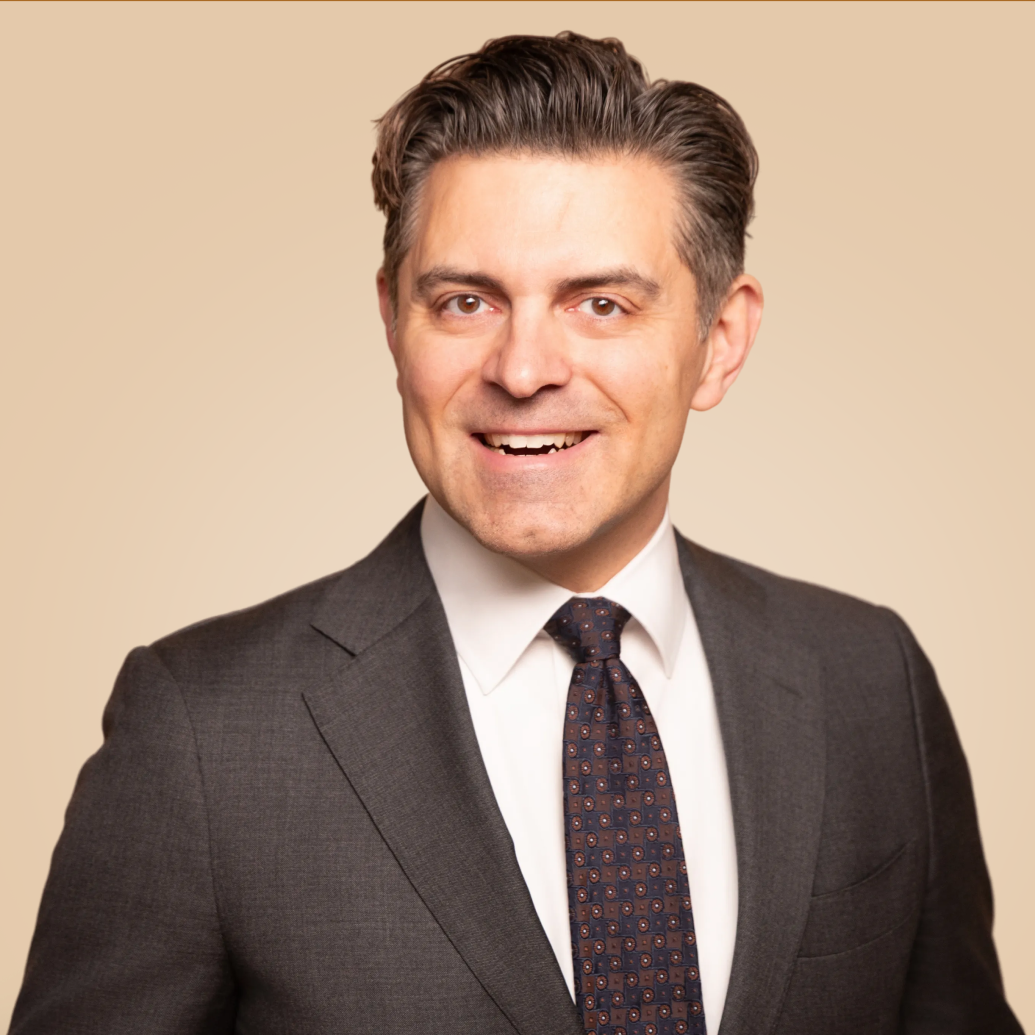 A smiling man wearing a dark gray suit, white shirt, and patterned tie, posing against a beige background.