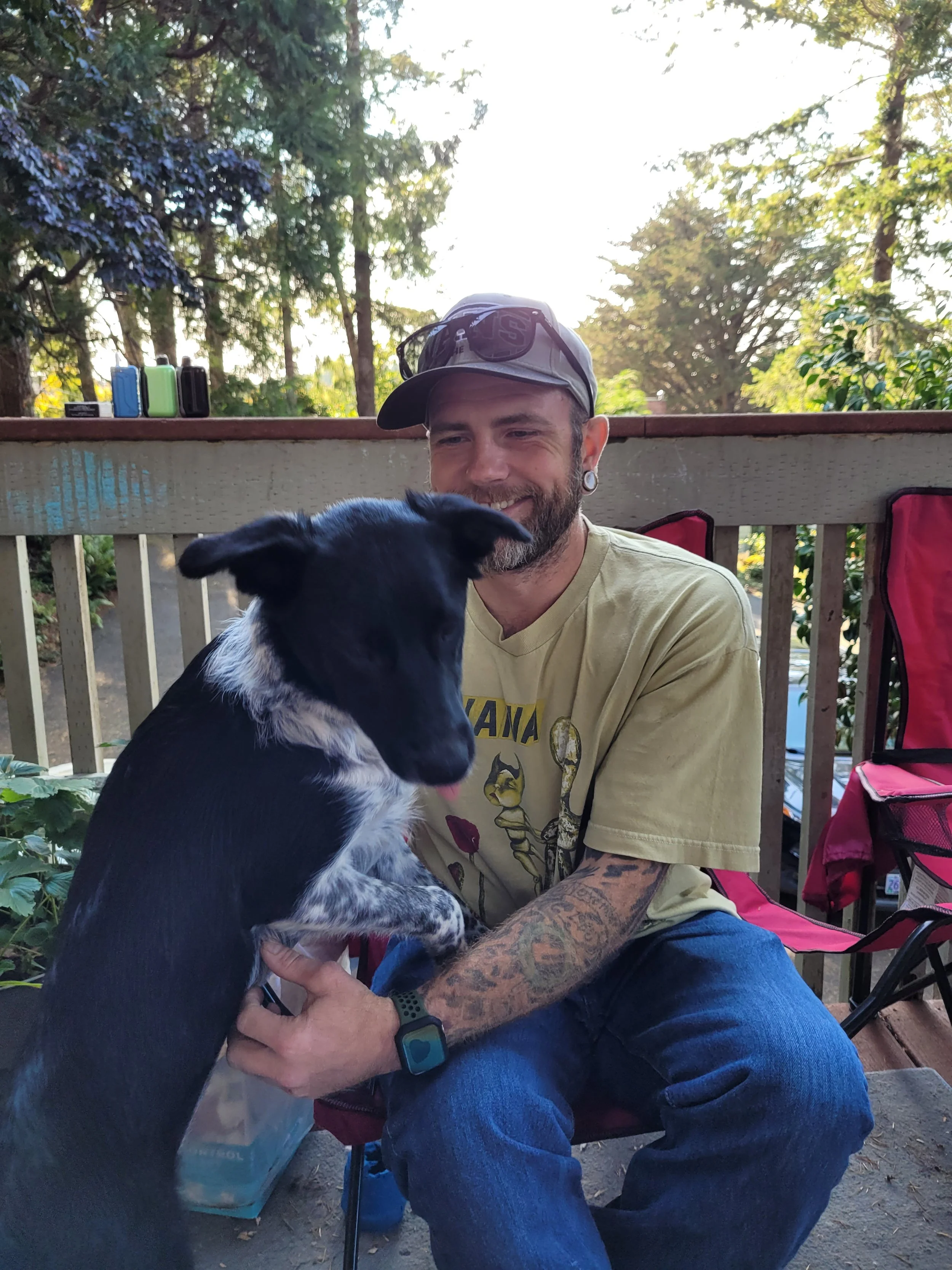 A man sitting on a chair on a porch, holding a black and white dog. Trees and outdoor furniture are visible in the background. Dune dog adventures Oregon coast dog daycare, Bandon dogs, Oregon coast dogs