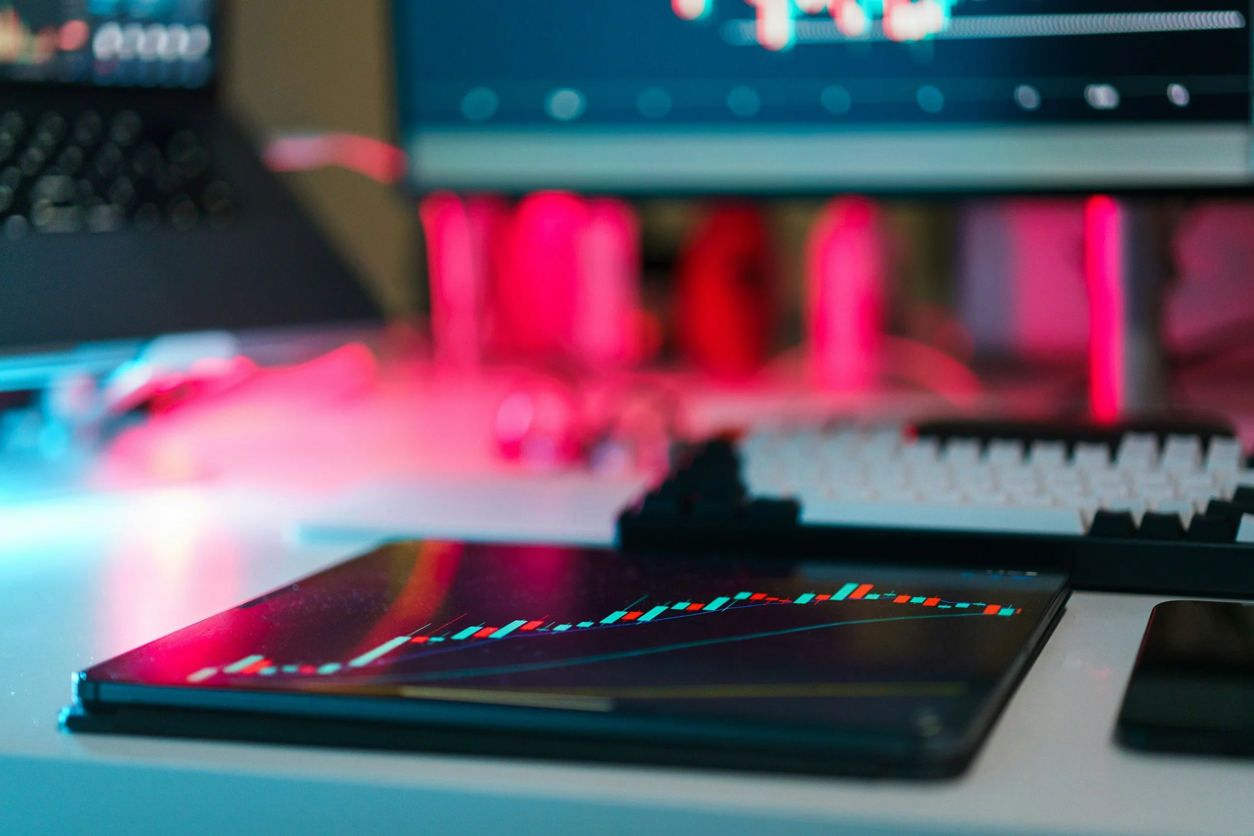 A tablet displaying a financial chart with candlesticks, placed on a desk with computer monitors and pink lighting in the background.