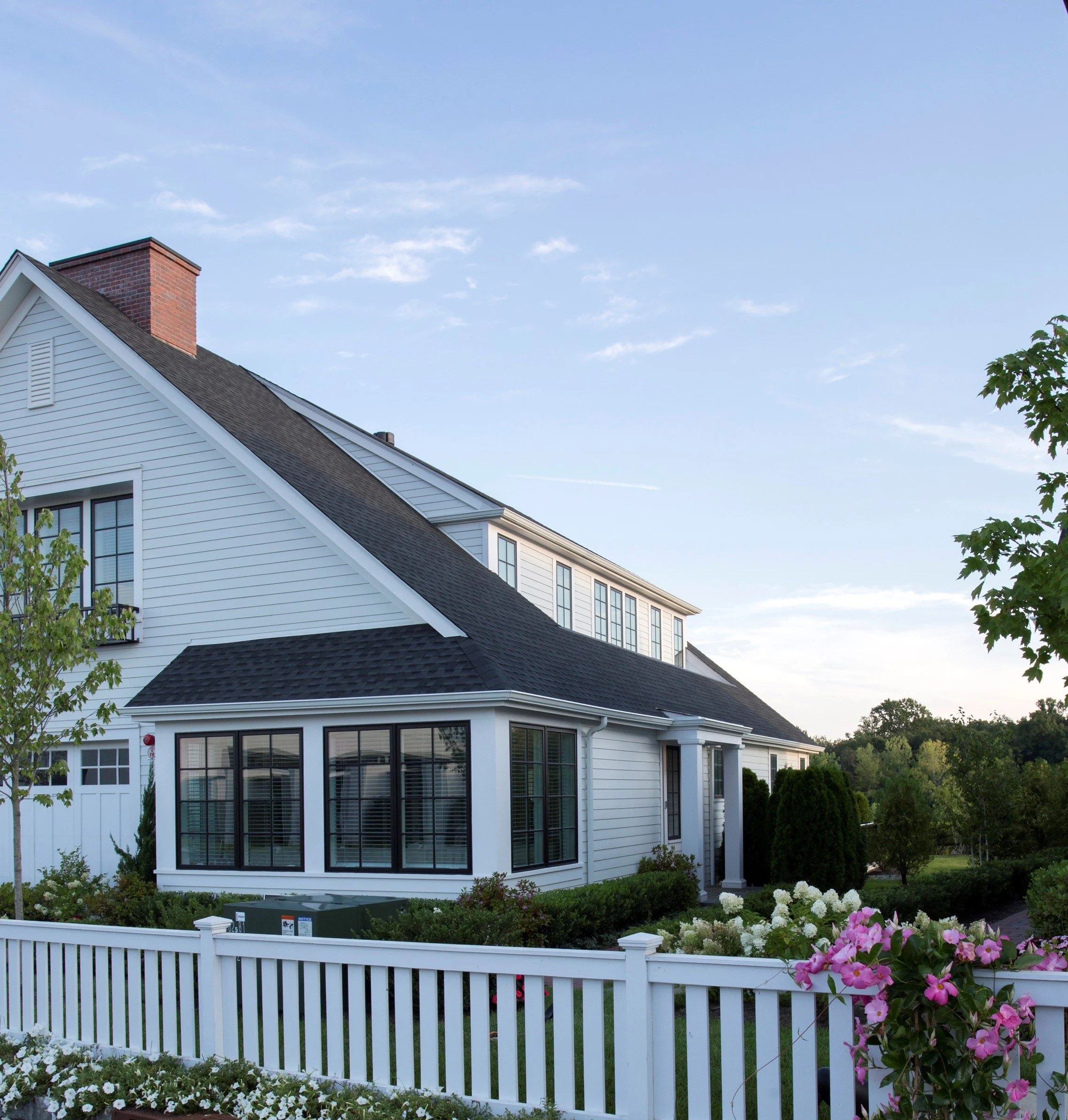 A two-story white house with black window frames, a black roof, a white picket fence, and well-maintained garden with pink and white flowers, green bushes, and trees under a blue sky.