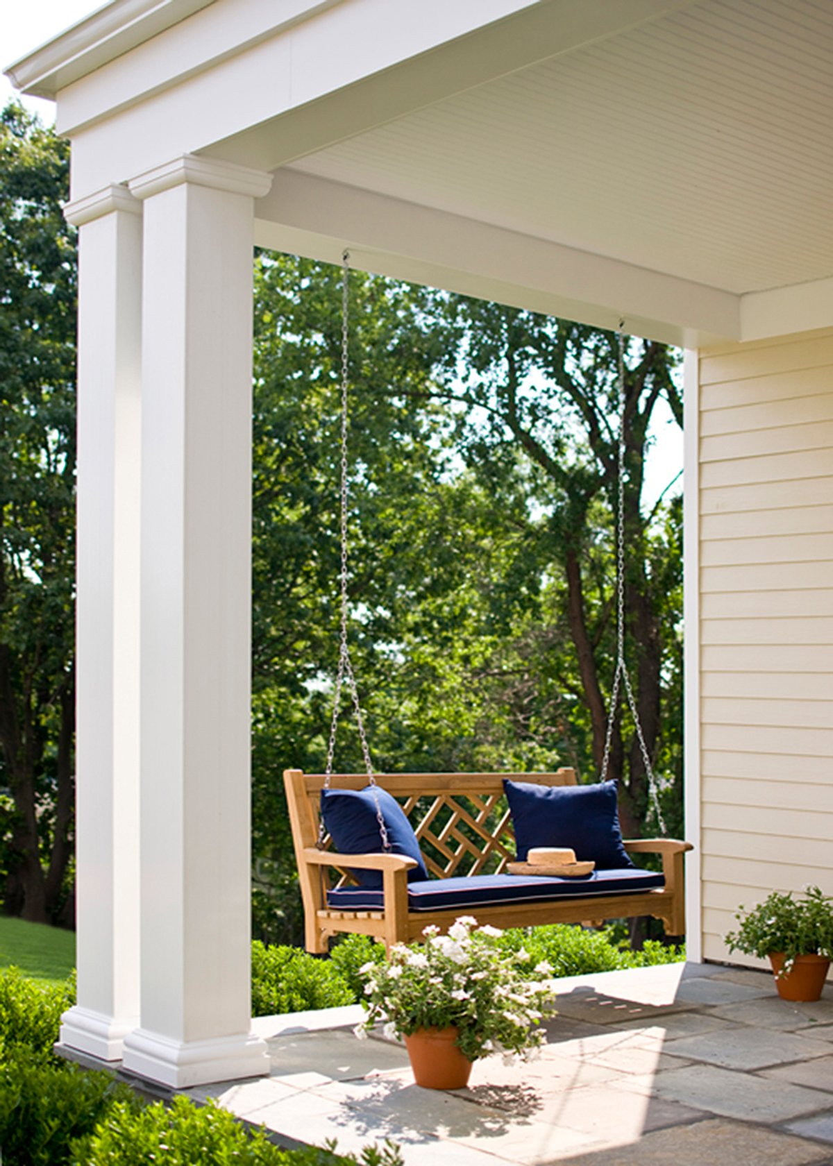 A porch with a wooden swing adorned with navy blue cushions, hanging from chains, and potted plants on a stone patio with greenery in the background.