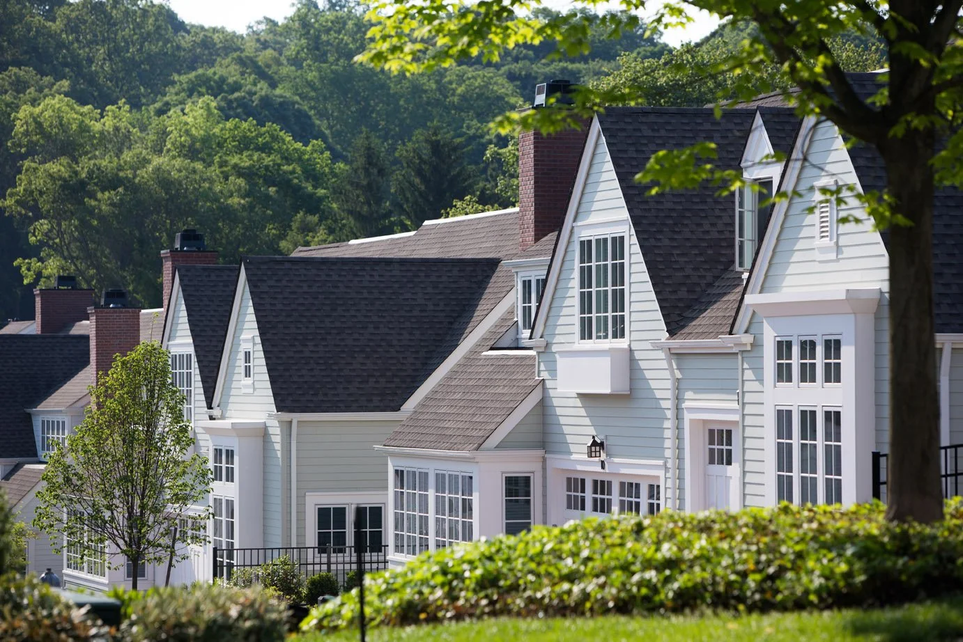 Row of white residential houses with steep dark roofs and large windows, surrounded by green trees and shrubbery.