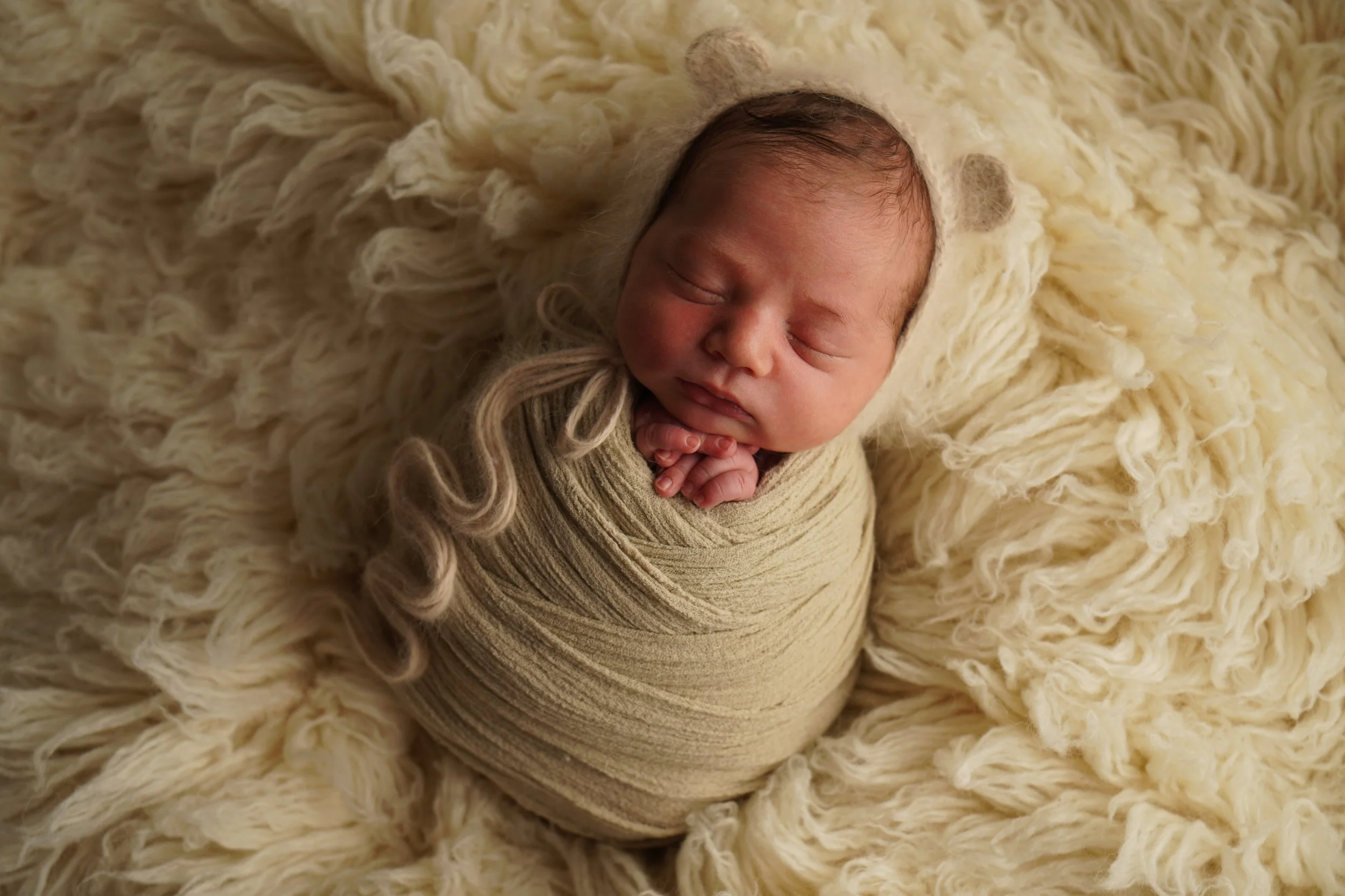 Ein neugeborenes Baby, eingepackt in einen beigen Stoff, liegt auf einem weichen, cremefarbenen Fell. Das Baby trägt eine passende Mütze mit kleinen Ohren und hat die Hände unter dem Kinn zusammengeklappt, während es schläft. Studiofotgrafie Rottweil
