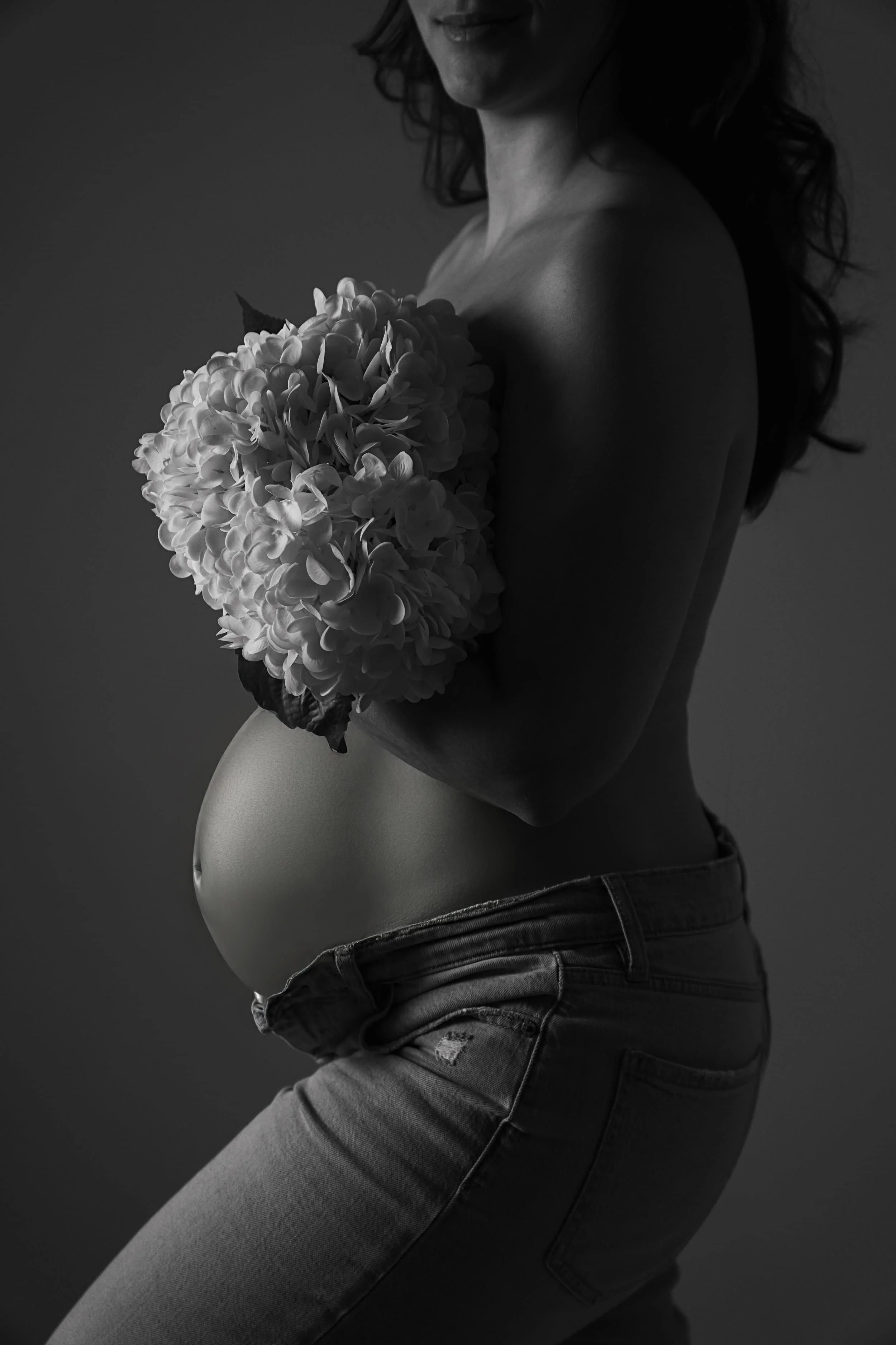 Schwangere Frau hält einen Blumenstrauß vor der Brust, lächelt leicht, trägt Jeans und hat lange, lockige Haare, in Schwarz-Weiß aufgenommen. KaysserArt Fotografie Rottweil