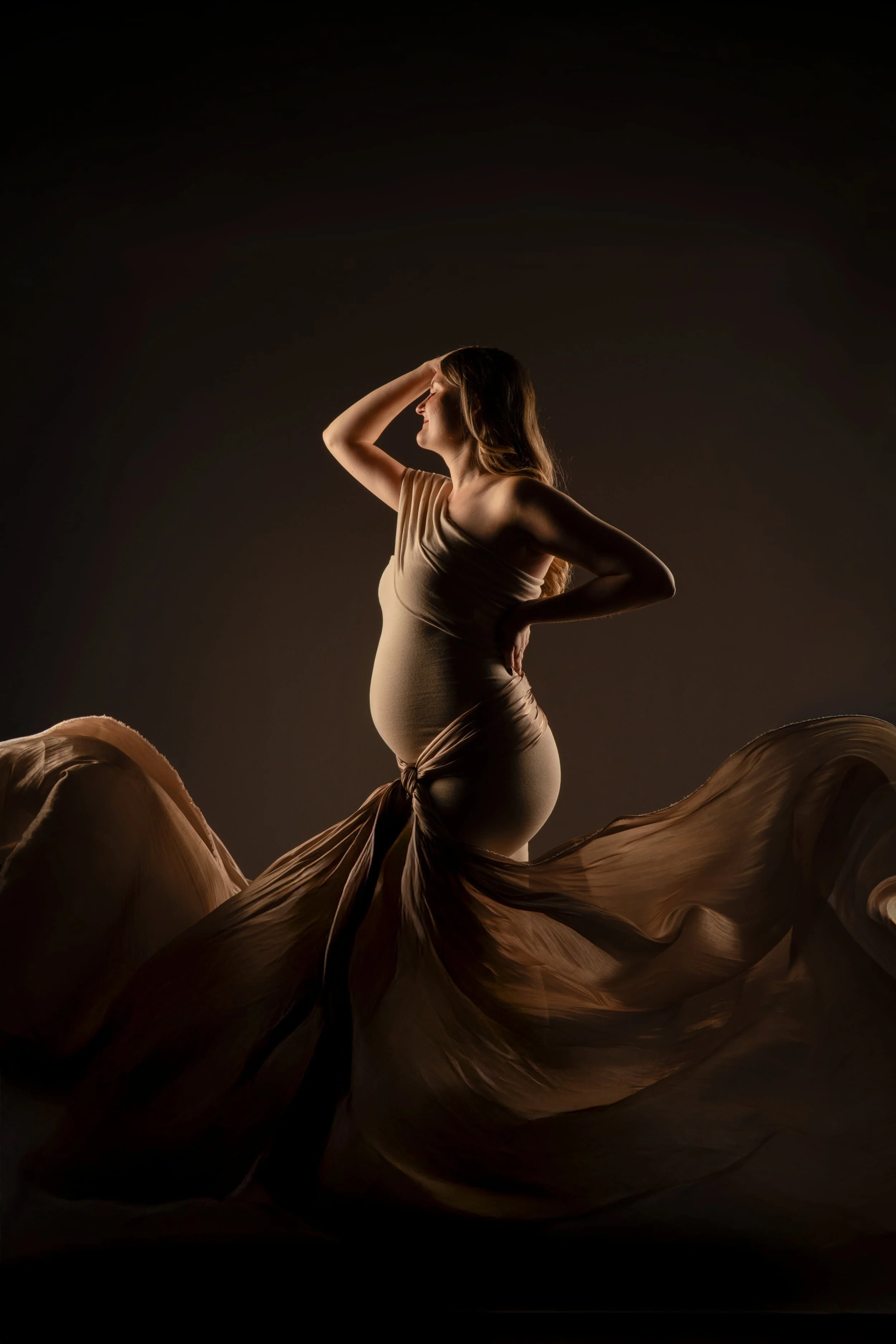 Eine schwangere Frau in einem fließenden, seidigen Kleid posiert vor dunklem Hintergrund, mit Hand im Haar, edler Stoff schwebt im Vordergrund und gibt dem Bild mehr Volumen und Tiefe. Studiofotografie von Robert Kaysser, KaysserArt Rottweil