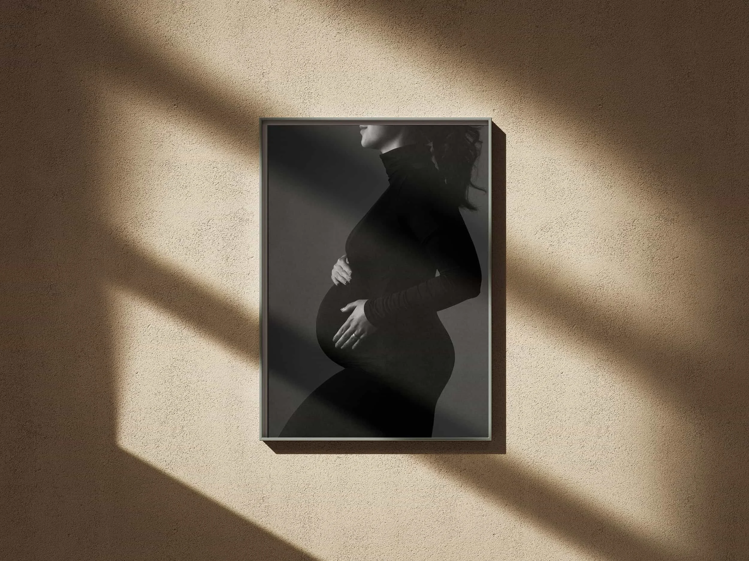 Schwarz-Weiß-Fotografie einer schwangeren Frau, deren Bauch sichtbar ist, in einem schwarzen engen Rollkragenkleid, auf einem dunklen Hintergrund, in einem Rahmen an einer beige Wand, mit diagonal verlaufenden Licht- und Schattenstreifen. KaysserArt
