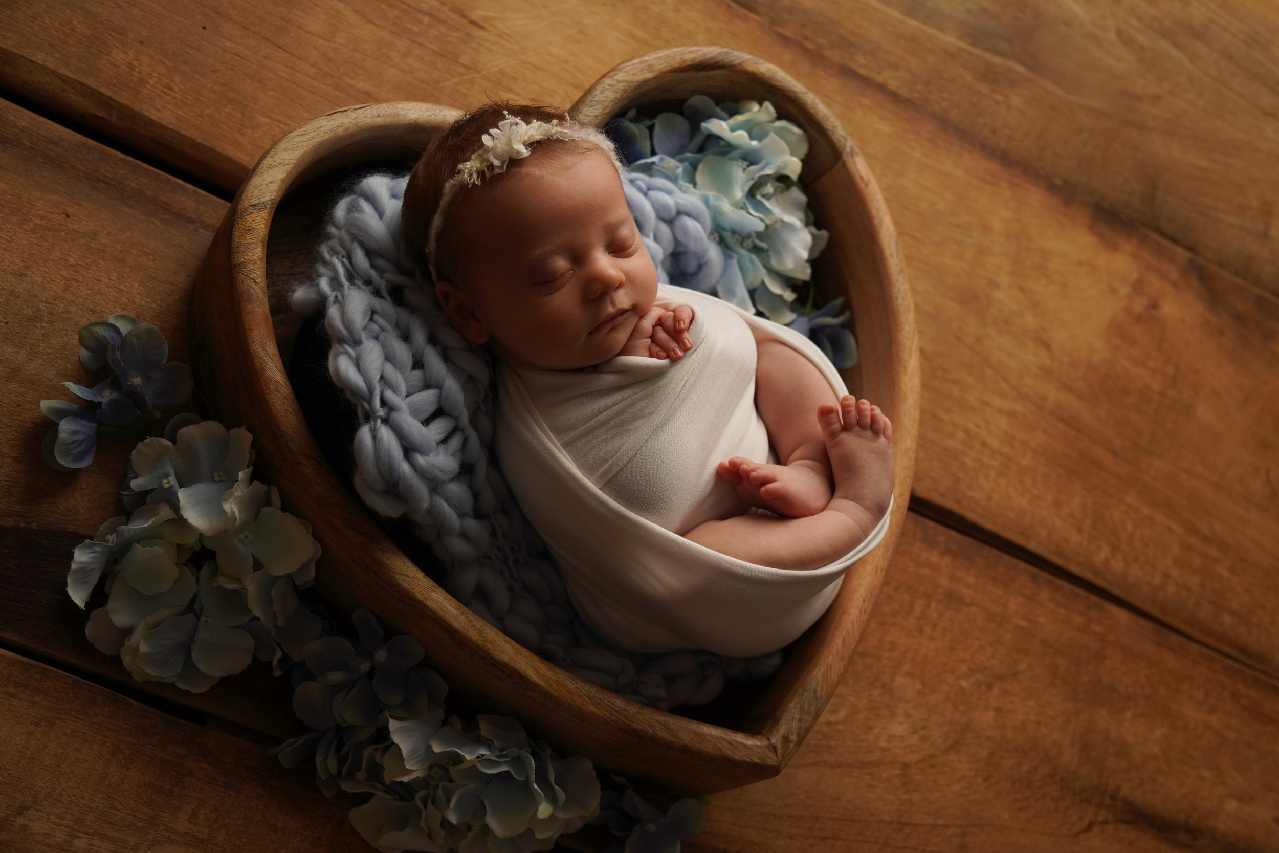 Neugeborenes Baby, eingewickelt in weiße Decke, liegt schlafend in einem Herz aus Holz auf einem Holzfußboden, umgeben von blauen und weißen Hortensien und einer weichen gehäkelten Decke. Studiofotografie von Robert Kaysser.