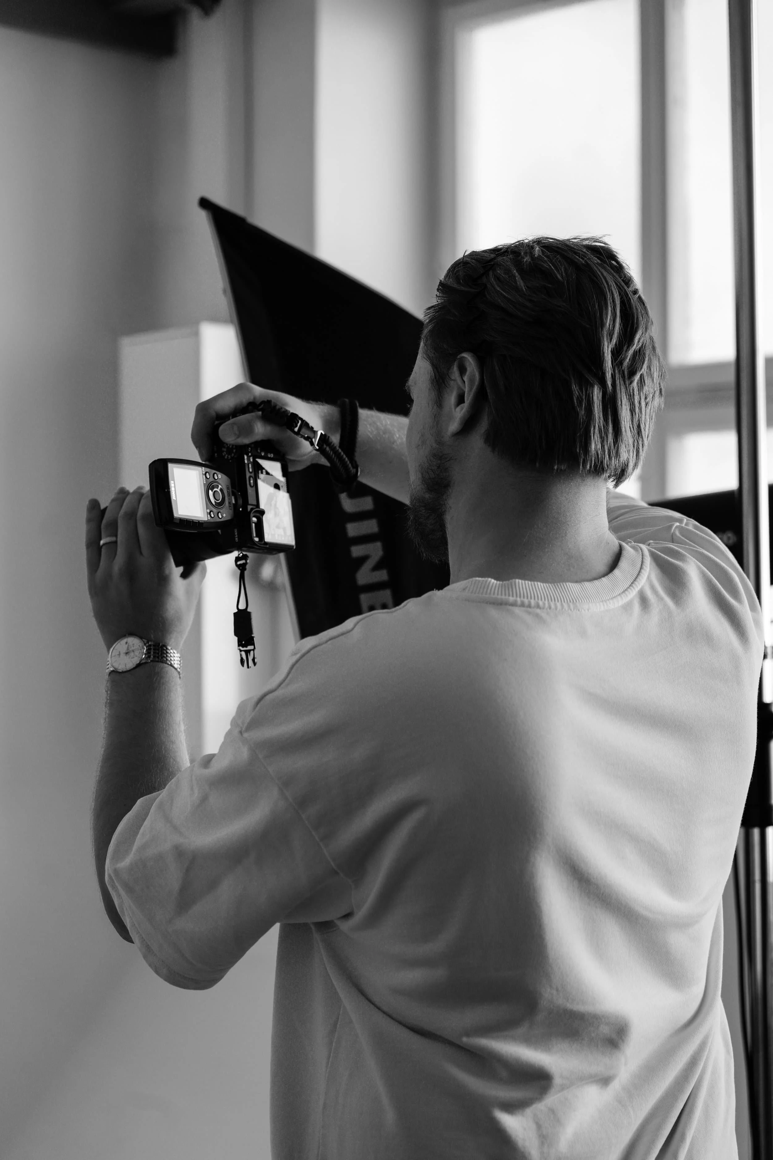 Ein Mann, Robert Kaysser, fotografiert mit einer Kamera in einem Studio, schwarz-weiß Aufnahme, Blick auf den Rücken und die Kamera, neben einem Scheinwerfer und einem Fenster.