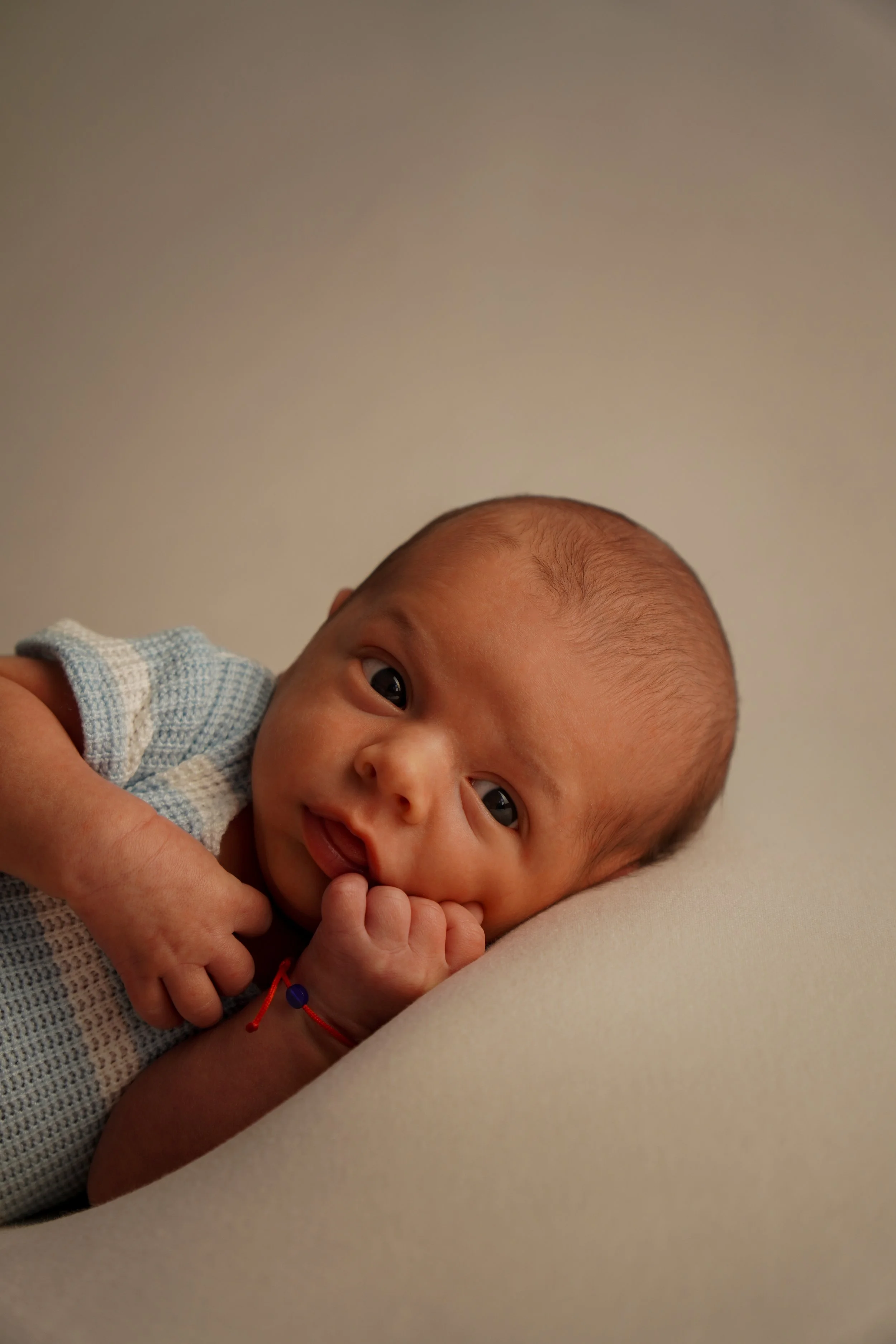 Ein neugeborenes Baby liegt auf der Seite, schaut neugierig in die Kamera, hat die Hand im Mund und trägt einen blau-weiß gestreiften, gestrickten Body, mit einem neutralen Hintergrund. Liebevolle Studiofotografie Schwenningen und Umgebung, Rottweil.
